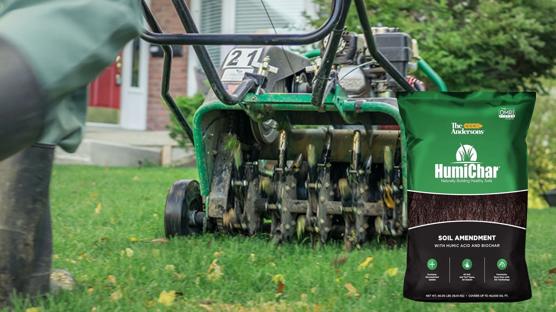 A green soil amendment bag labeled 'Humichar' with black and white text, placed in front of a lawn aerator operating on grass. The background shows a house with brick and siding, and trees.