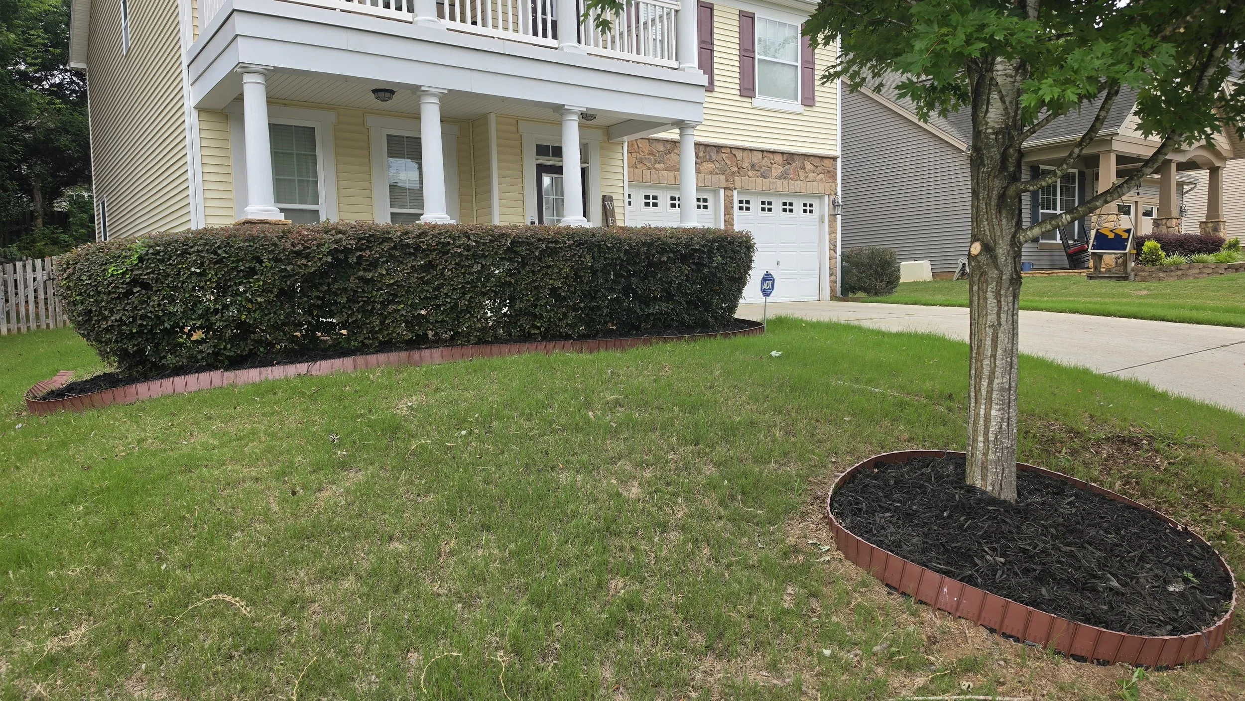 A front yard with a trimmed shrub and a newly planted tree surrounded by a curved plant bed, with a house featuring a covered porch, white columns, yellow siding, and a garage in the background.