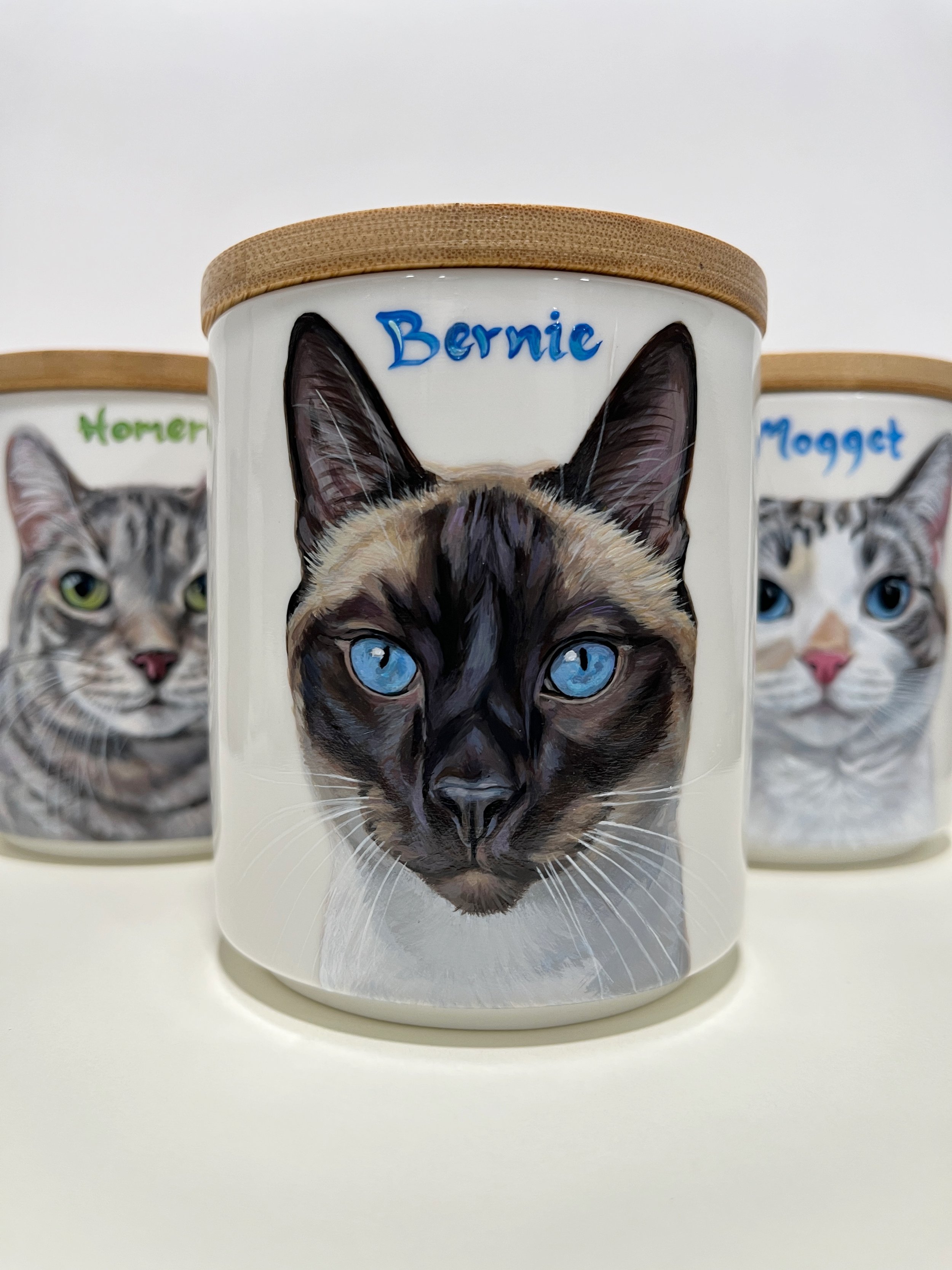 photo of three cat portrait urns they are hand painted portraits on ceramic urns with wood lids. the cat in the front is named Bernie his name Is painted in the same color as his eyes. this link takes you to commission shop page