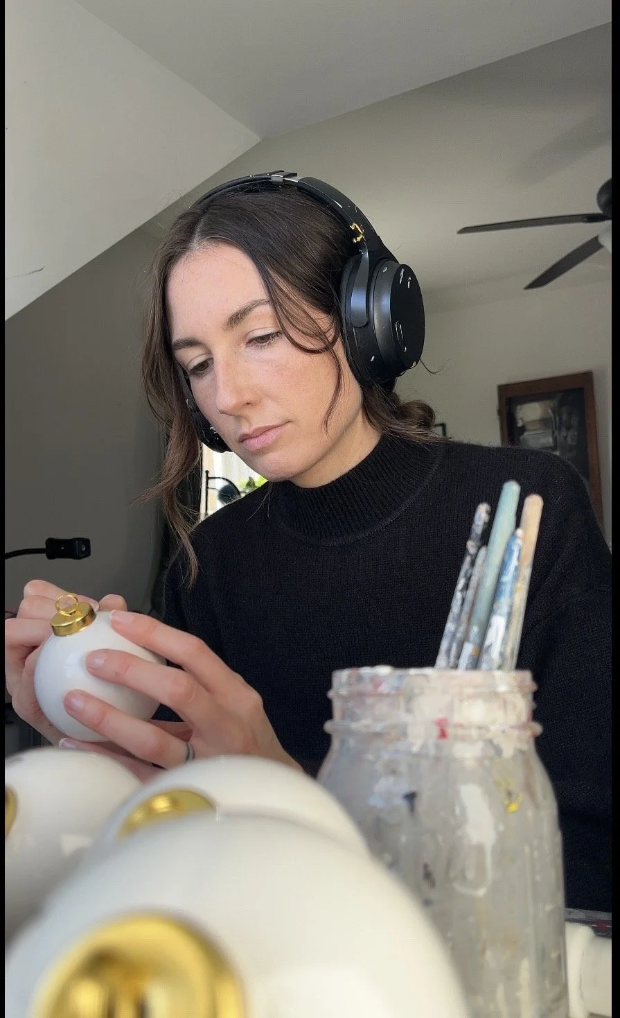 photo of the artist painting at drafting table she  has brown hair and is wearing a black sweater along with black headphones. there is an ornament in her hand and a jar of paint brushed in the foreground