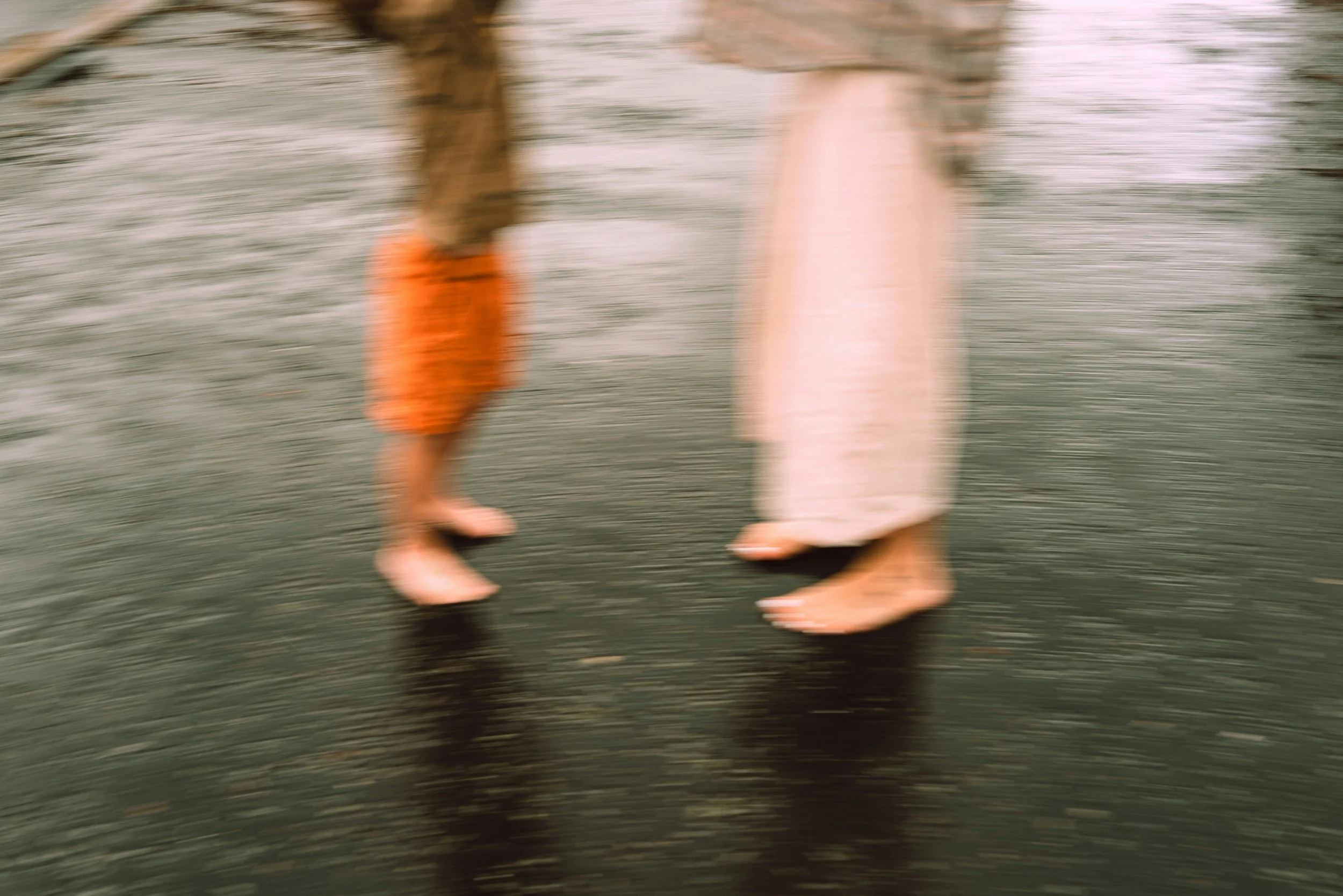 Blurred image of two people walking barefoot on a beach, one wearing orange pants and the other in cream-colored pants.
