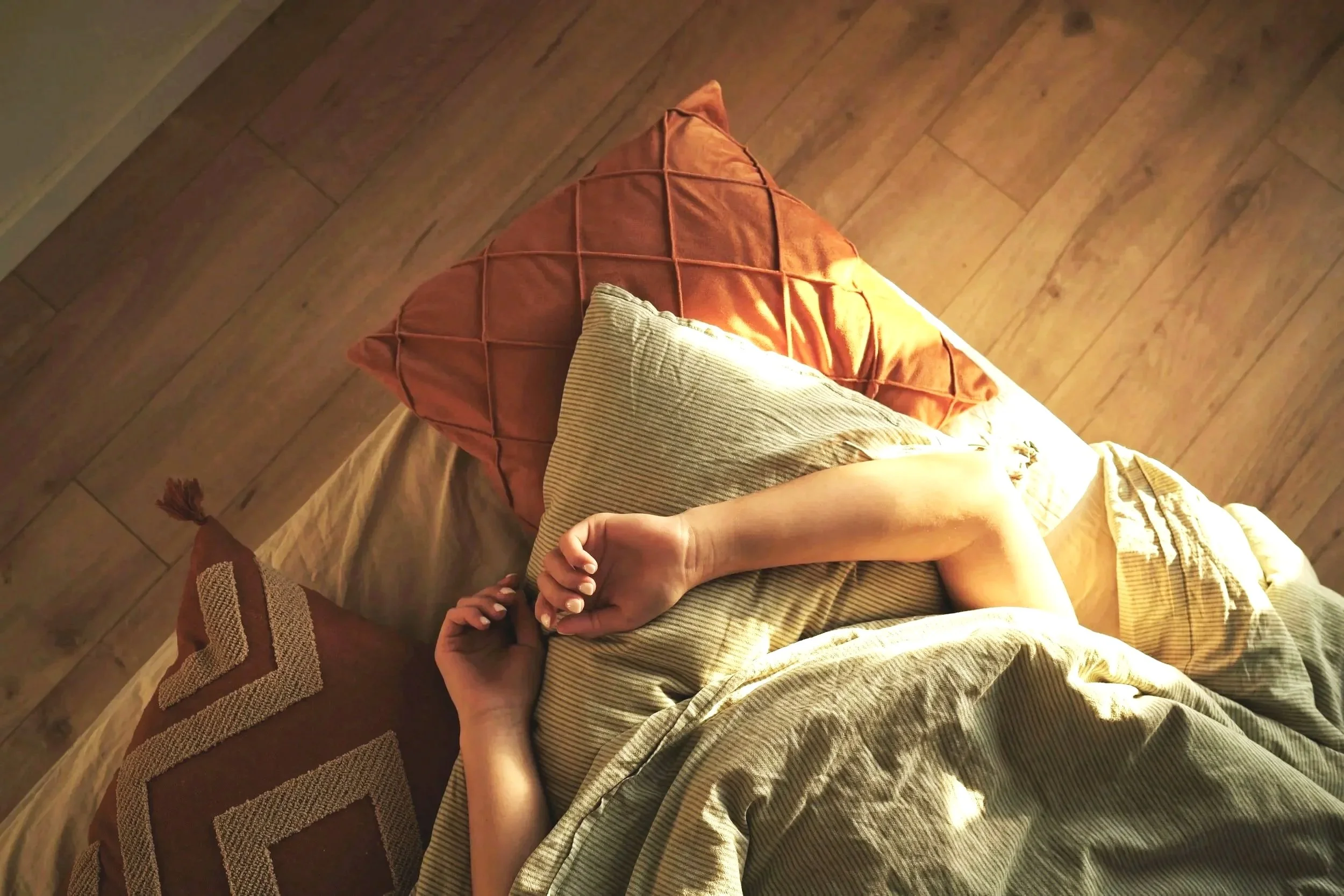 View of a person's arm resting on their lap, with their hand holding another person's hand. The scene is set indoors on a wooden floor, featuring a carrot-colored pillow and a striped beige and white blanket or cloth.