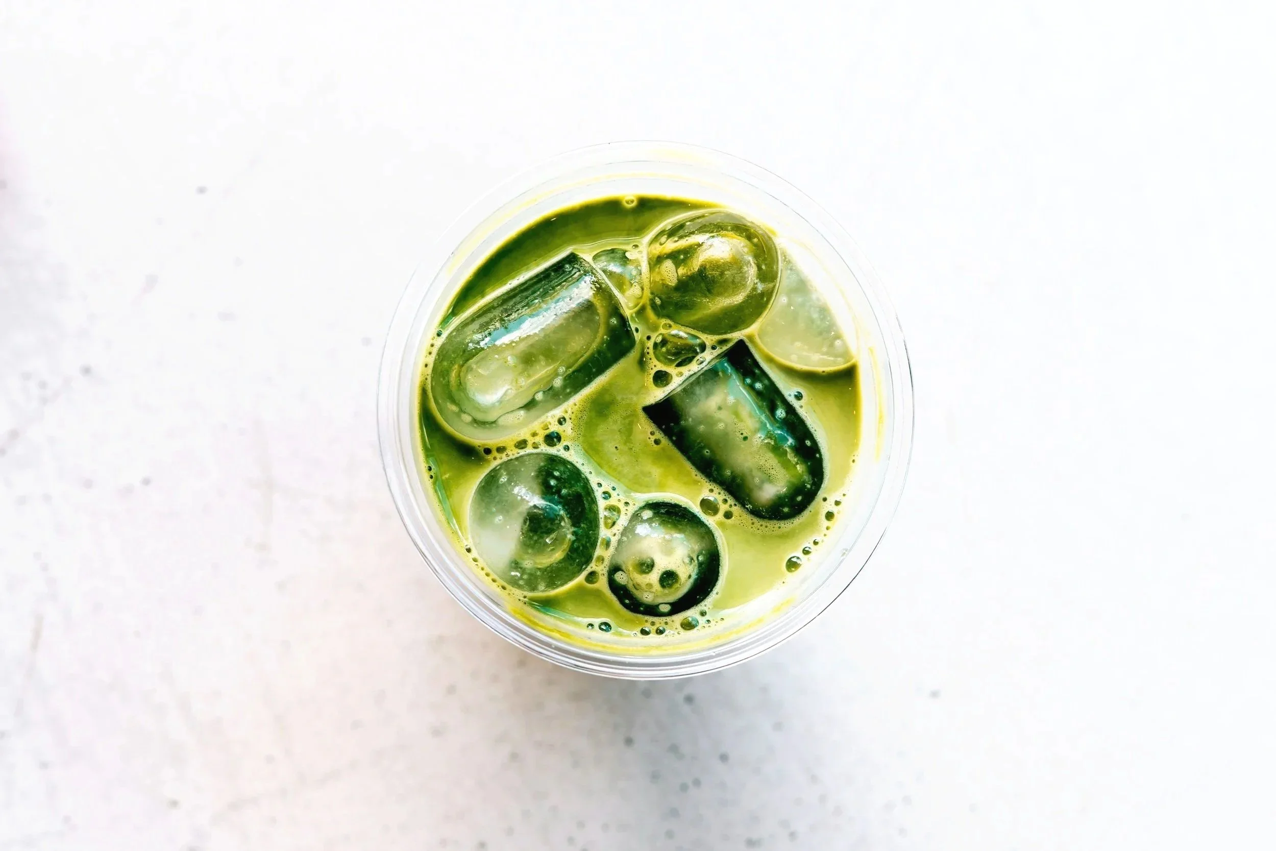 A clear plastic cup filled with green liquid and ice cubes, placed on a white surface.