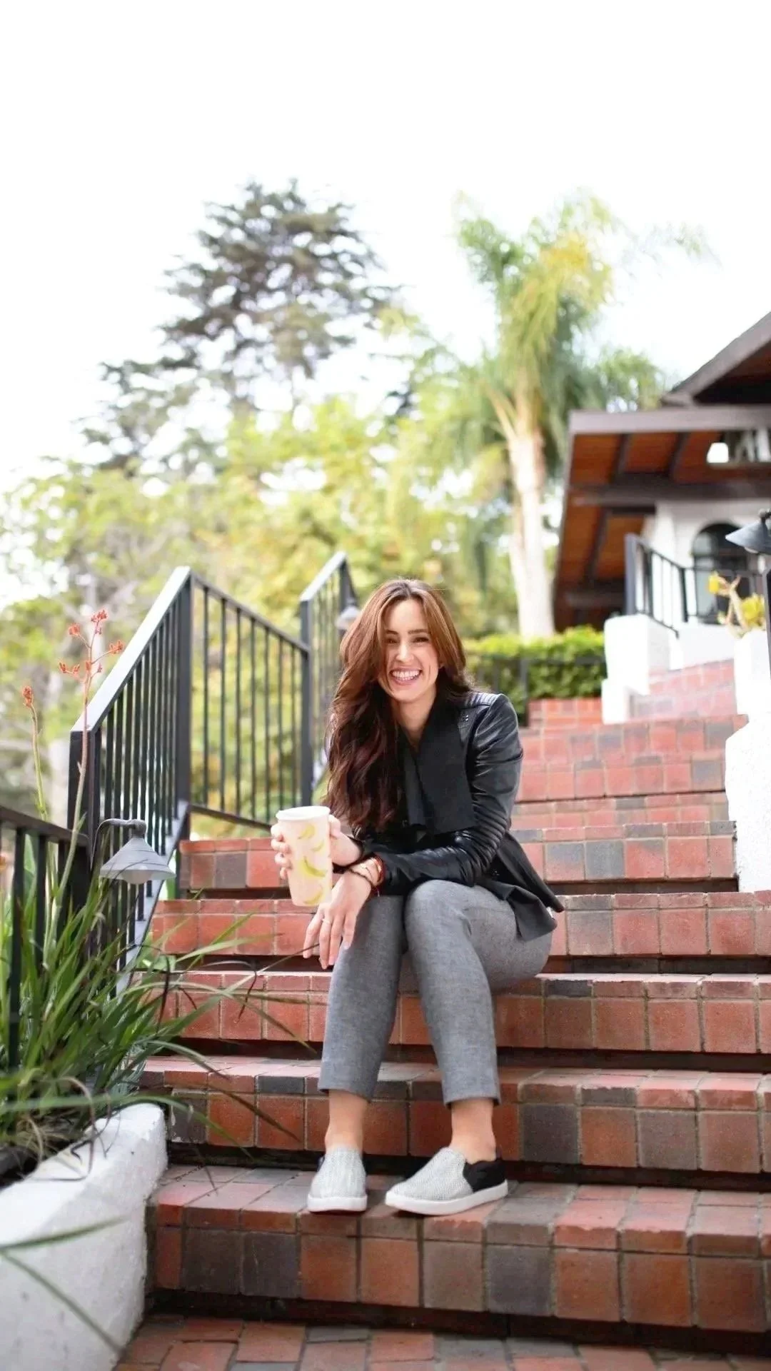 A woman with long brown hair, wearing a black jacket and gray pants, sits on brick stairs outdoors smiling while holding a disposable coffee cup.
