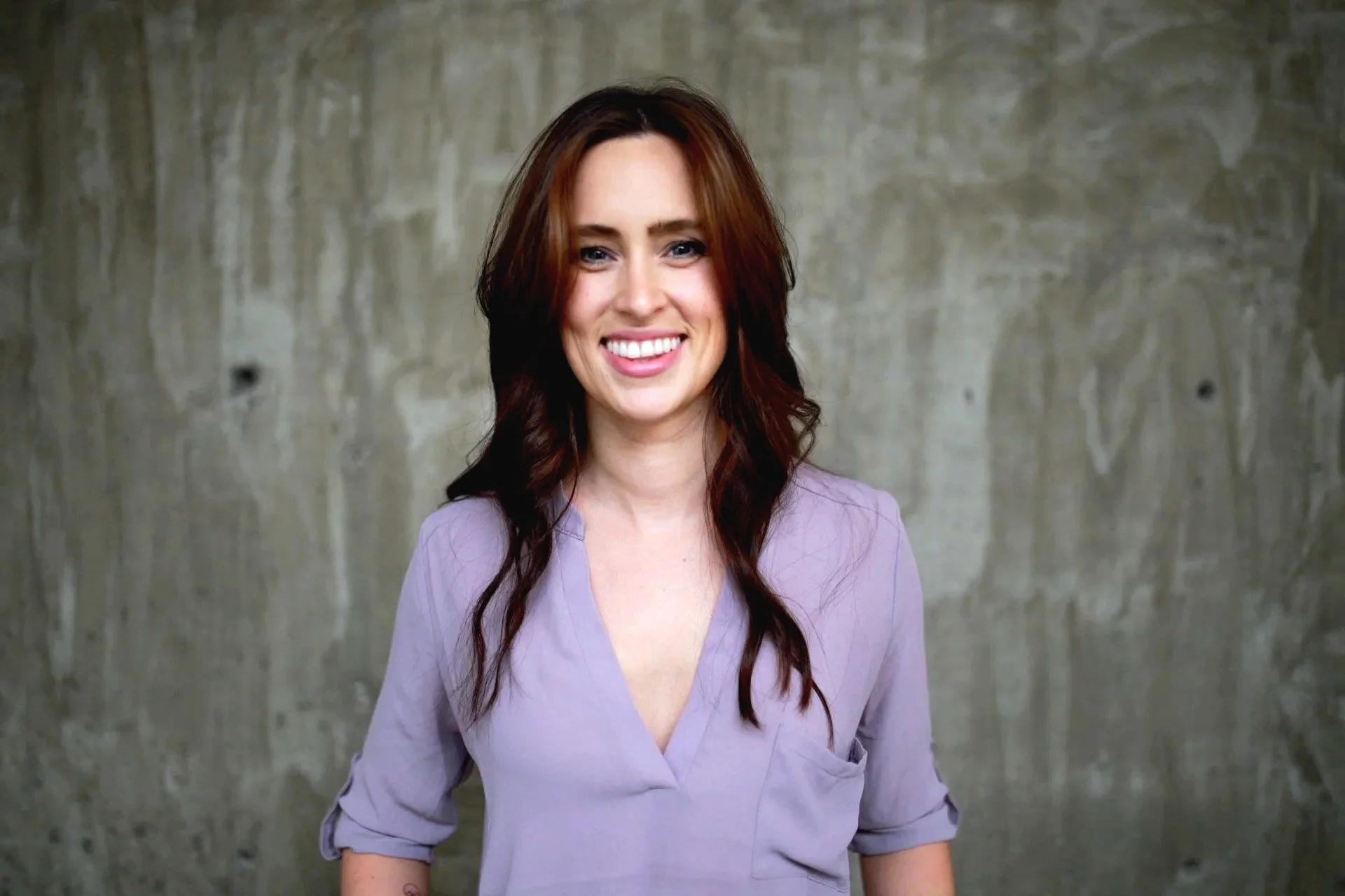 A woman with long, wavy brown hair and a bright smile standing against a textured concrete wall, wearing a light purple blouse with rolled-up sleeves.