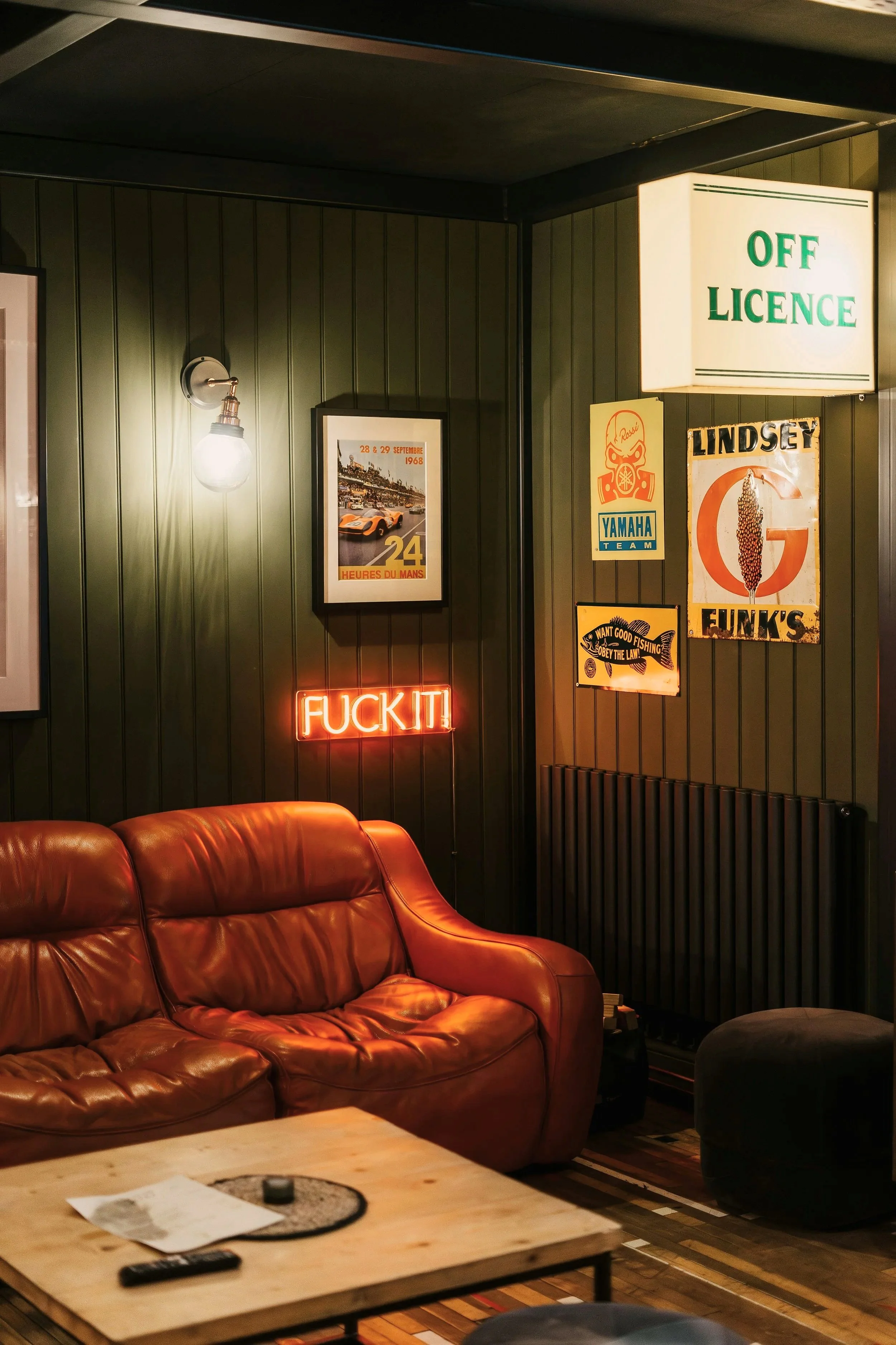 A cozy indoor setting with a worn leather sofa, a coffee table with papers, a lighter, and a TV remote, and walls decorated with various posters and signs including a neon sign that reads 'FUCK IT' and a bright backlit sign that says 'OFF LICENSE'.
