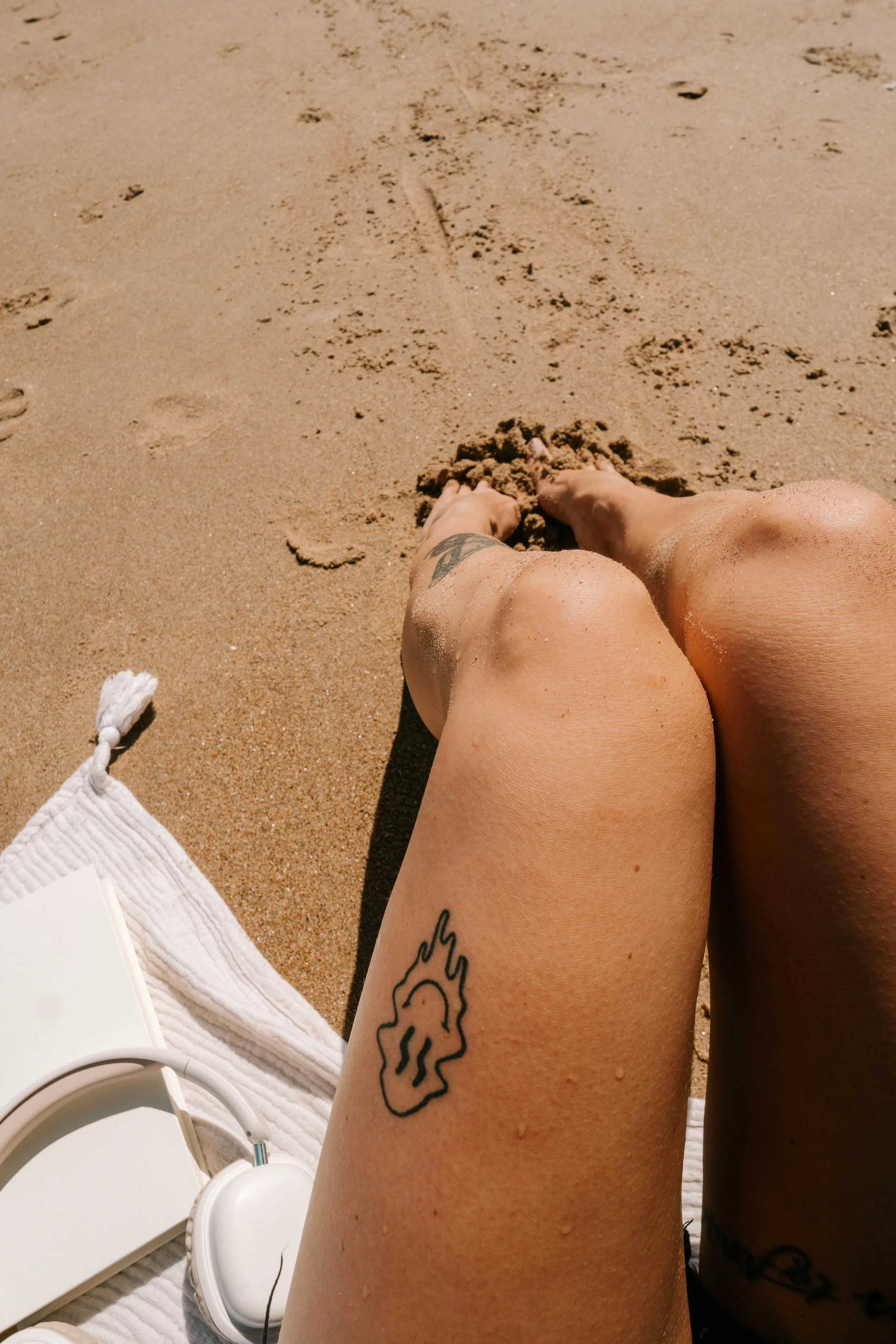 A pair of tanned legs with water droplets, one tattoo of a lion and a white towel, headphones, and a white book or paper on a sandy beach.