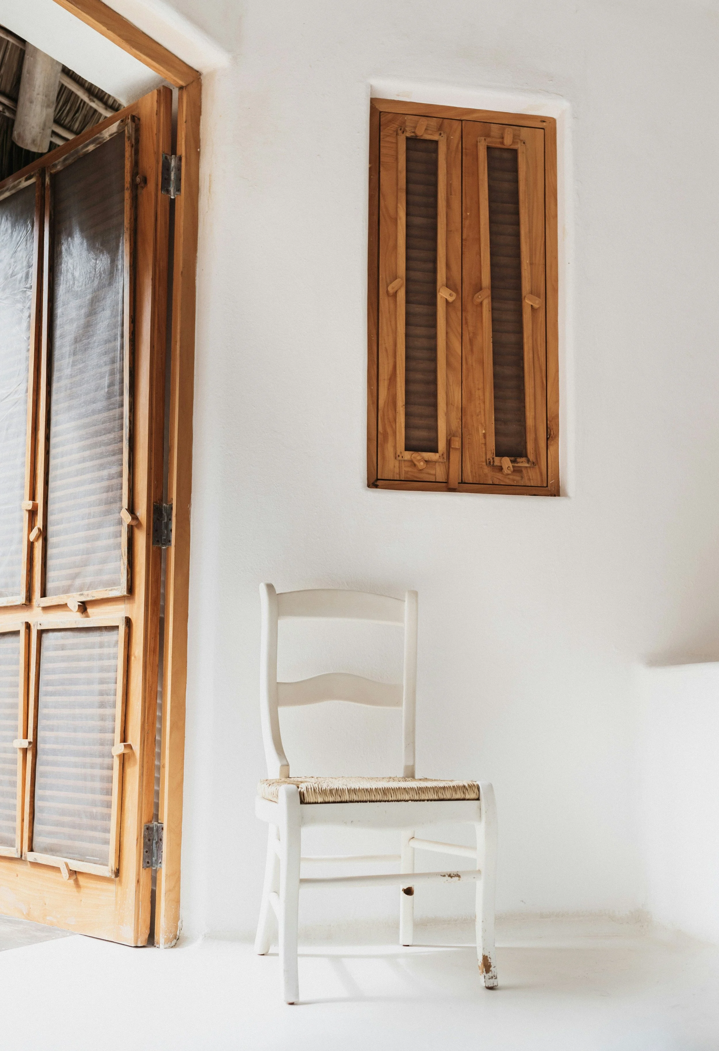 A rustic white interior with a wooden window with louvered shutters, a wooden door with glass panes, and a white wooden chair with woven seat.