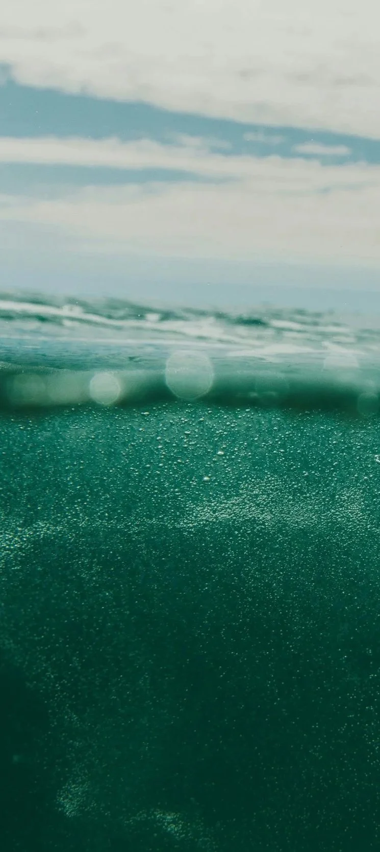 A view of the ocean with a clear blue sky and clouds above, seen from below the water surface, showing bubbles and water particles.