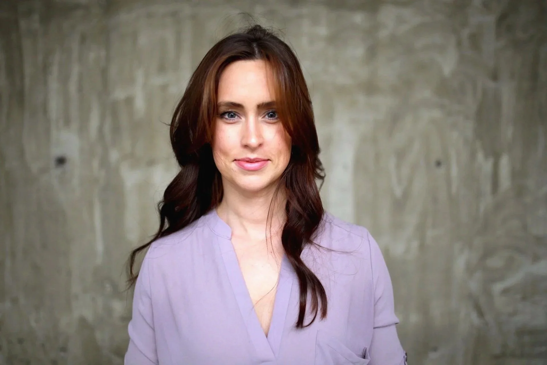A woman with long, dark red hair, blue eyes, and fair skin stands in front of a plain concrete wall, smiling softly. She is wearing a light purple blouse with a V-neck.