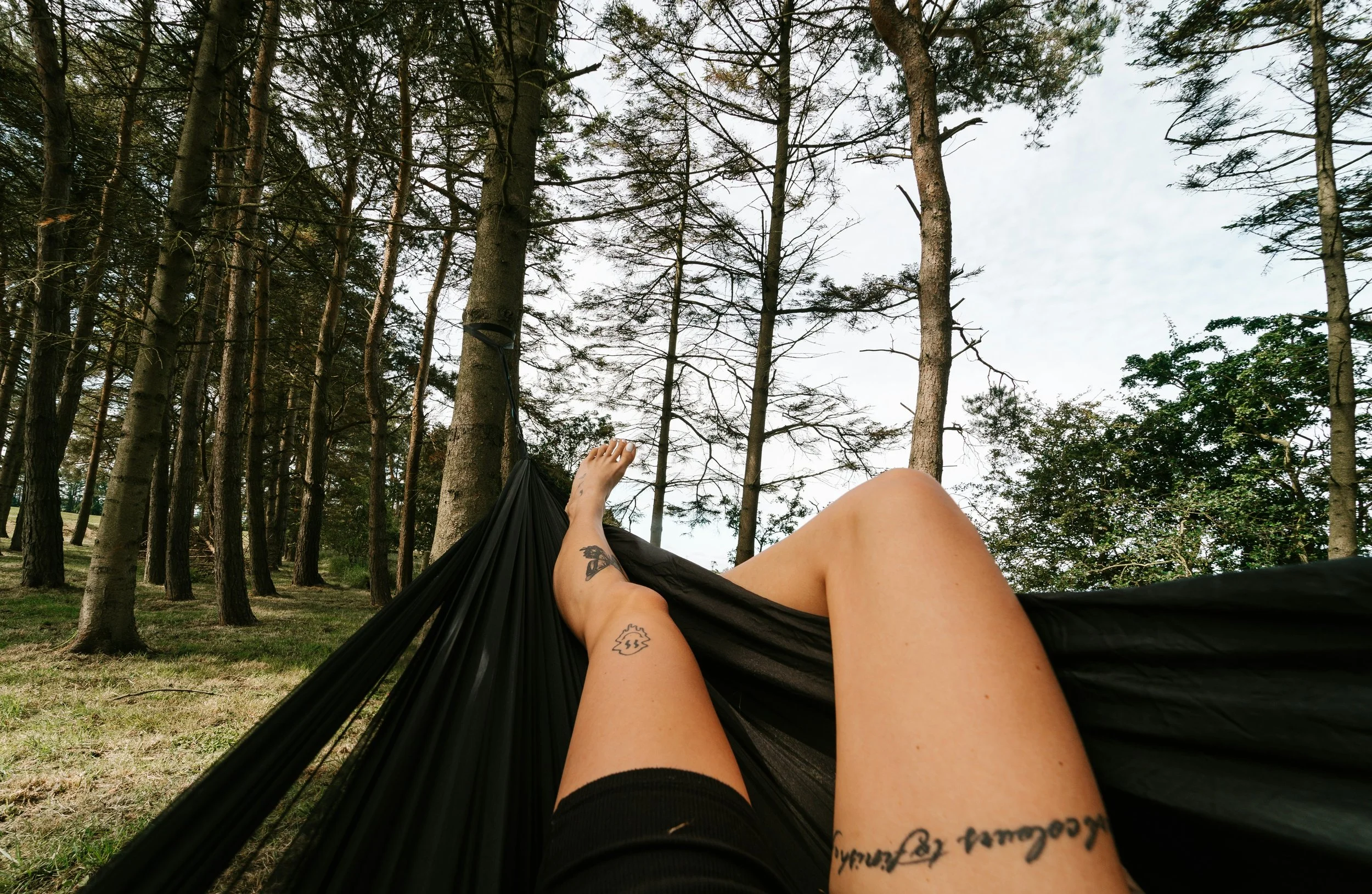 Person lying in a black hammock in a forest, with their legs and feet visible, surrounded by tall trees and a partly cloudy sky.