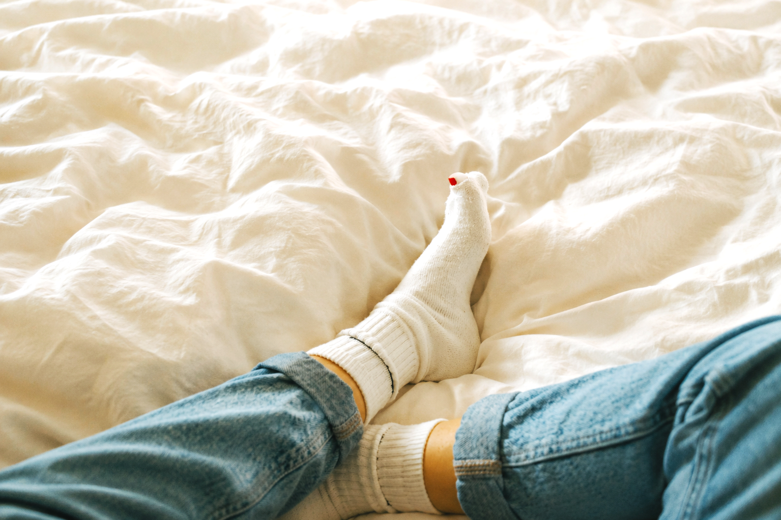 A person's legs and feet in blue jeans and white socks are resting on a rumpled white blanket or bedspread.