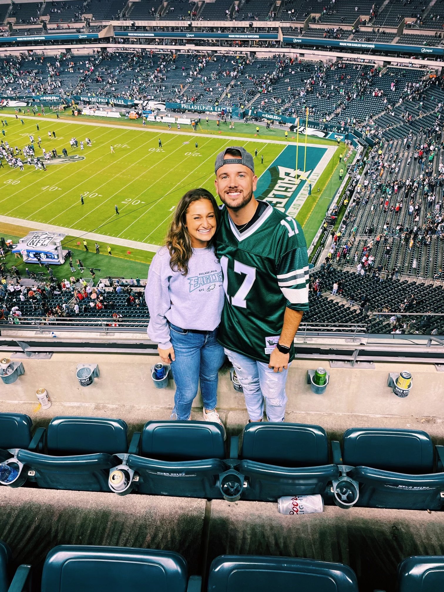 Two people smiling and standing together in a sports stadium, with a football field and spectators in the background. One is a woman with long hair wearing a gray Philadelphia Eagles sweatshirt, and the other is a man wearing a Philadelphia Eagles jersey and a backward gray cap.