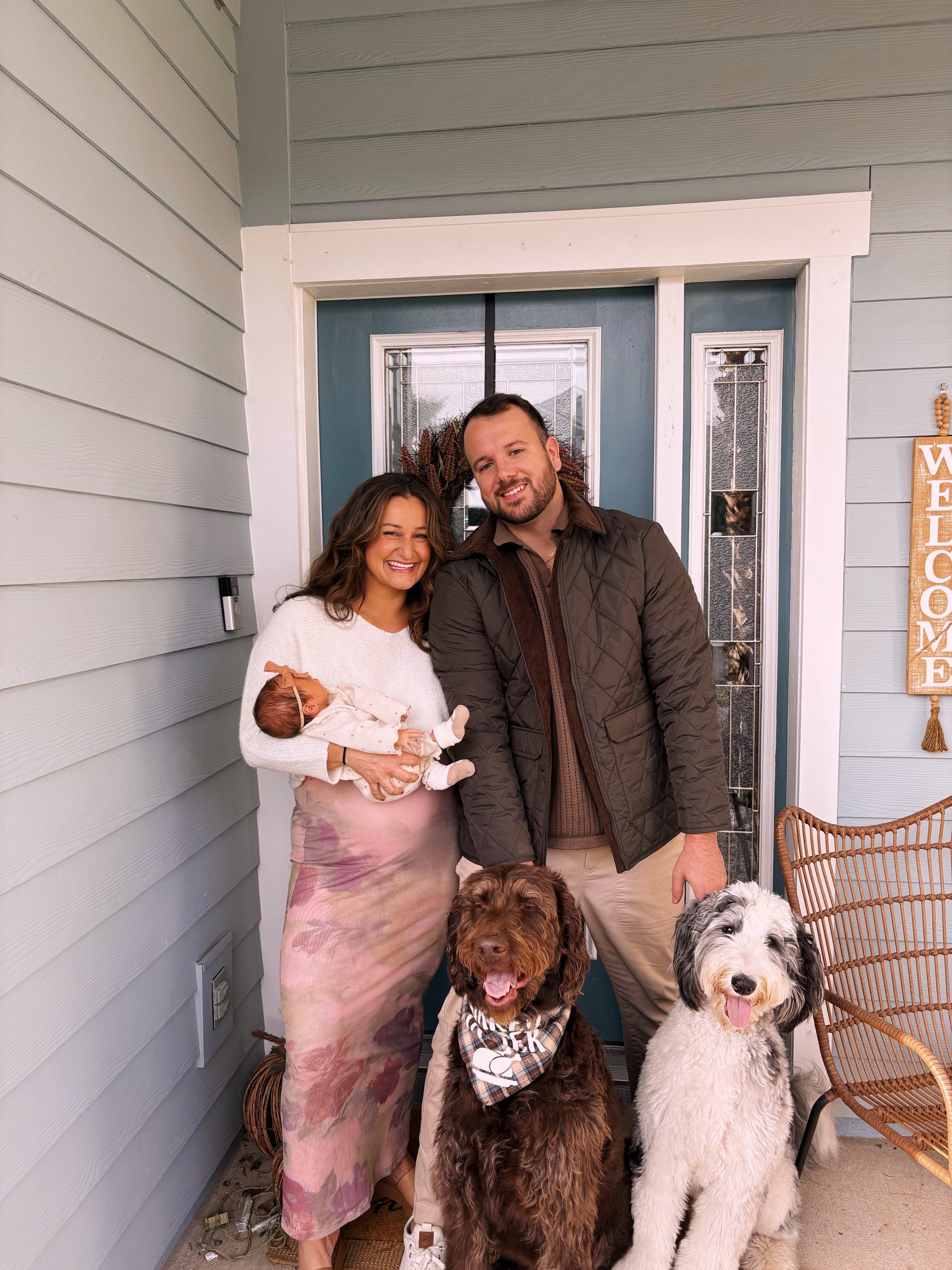 A woman holding a newborn baby and a man standing on a porch next to two dogs, with a decorated front door in the background.