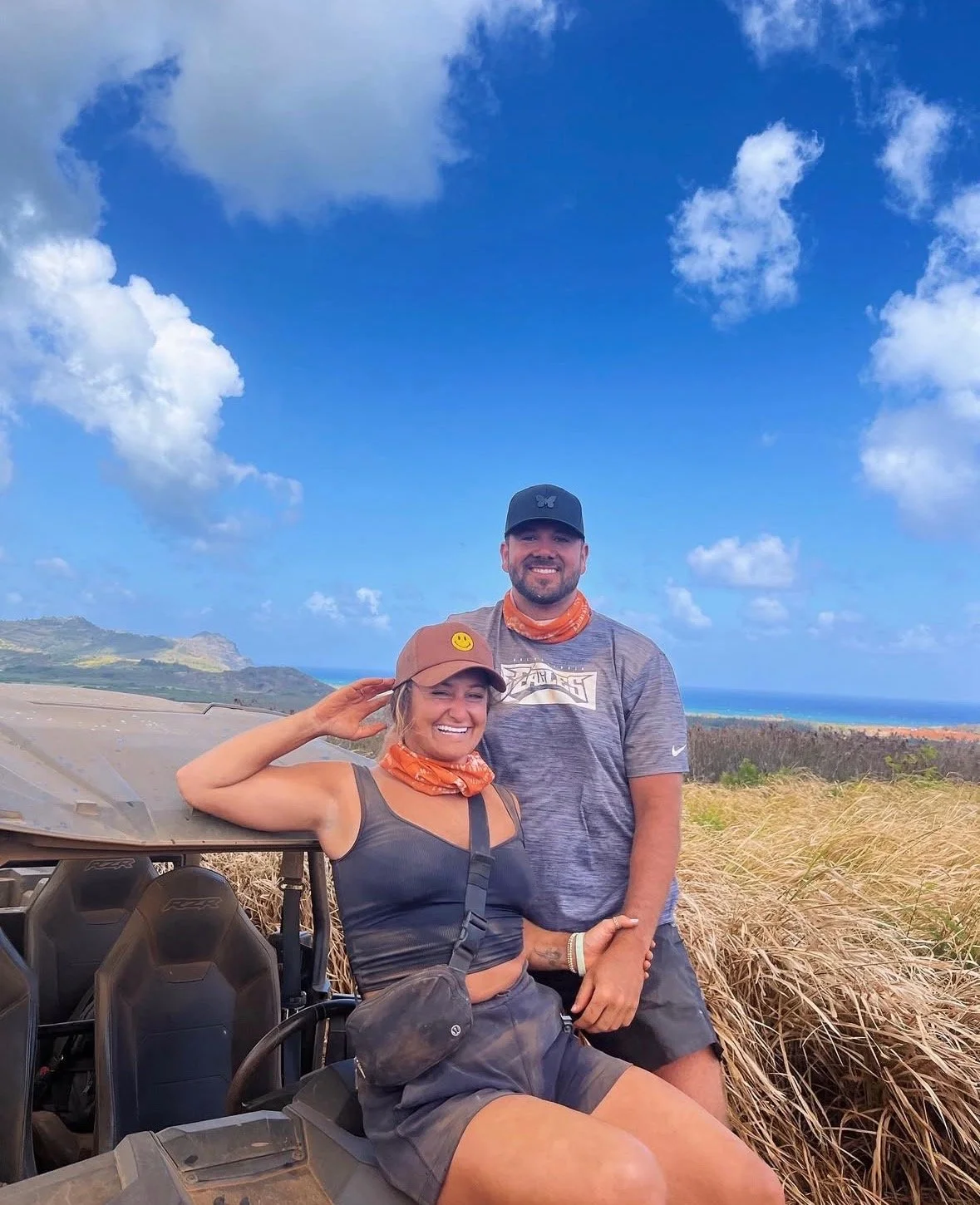 A smiling man and woman outdoors in a field with tall grass, with hills and a cloudy blue sky in the background. The woman is sitting on a vehicle with off-road tires, wearing a brown cap with a smiley face pin, and an orange bandana around her neck. The man is standing behind her, wearing a black cap, a gray T-shirt, and an orange bandana around his neck.