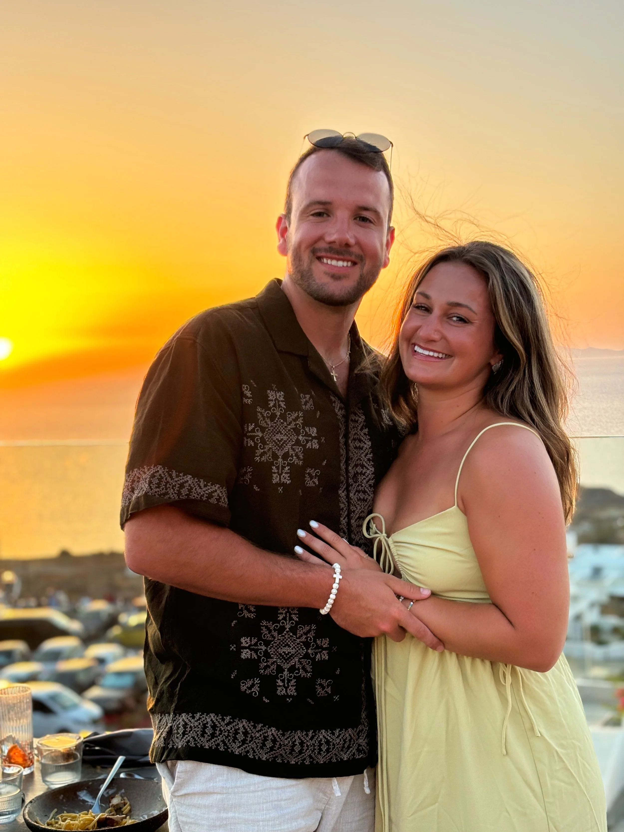 A young couple with light skin smiling and embracing at sunset by the water, with parked cars and outdoor tables in the background.