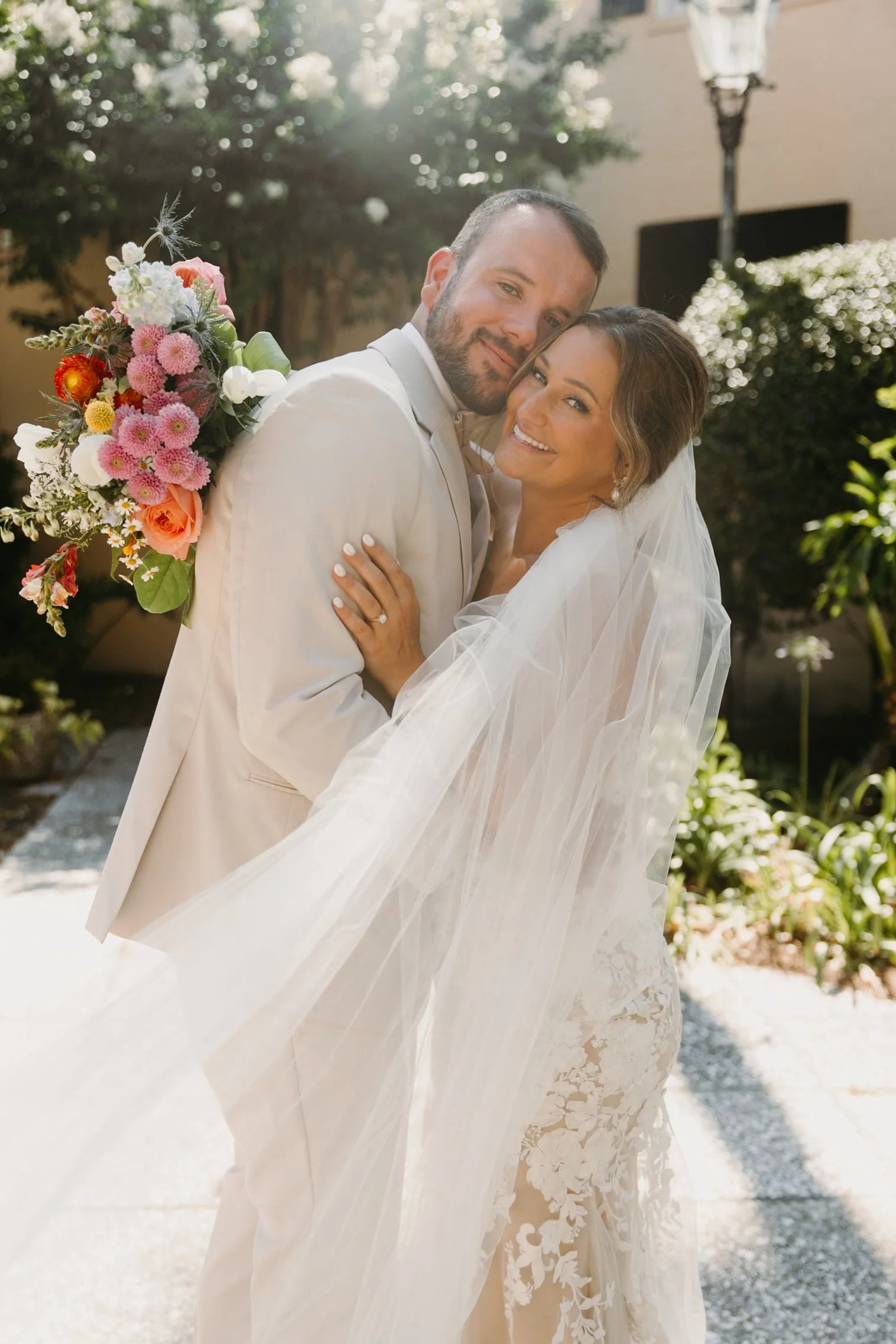 A bride and groom smiling and embracing outdoors, with the bride wearing a lace wedding dress and holding a bouquet of pink, white, and orange flowers. The groom is wearing a light-colored suit. Sunlight filters through trees in the background.
