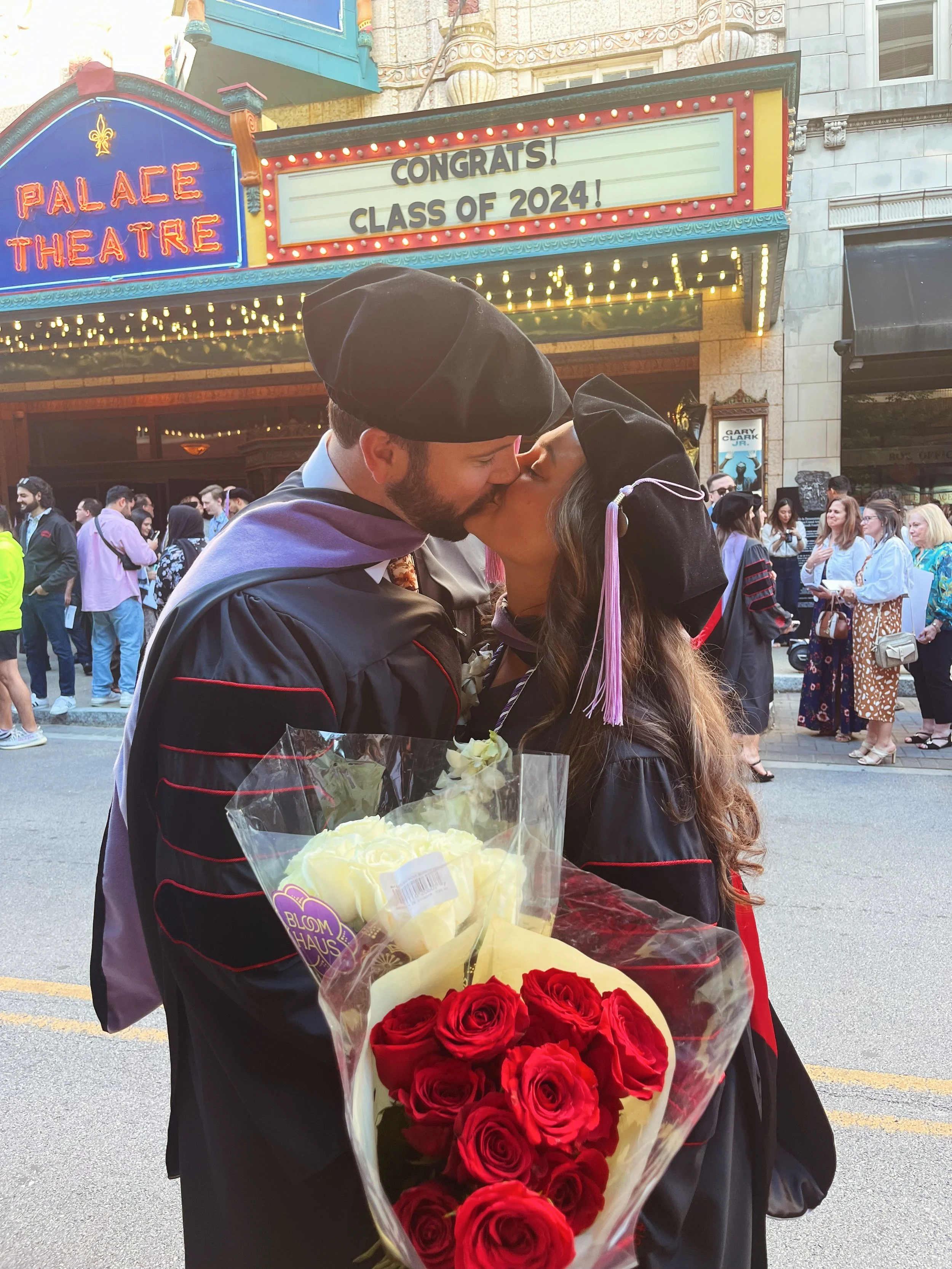 A man and woman in graduation gowns and caps kiss at a graduation ceremony. The woman holds a bouquet of red roses. People gather and take photos in the background, with a marquee sign displaying congratulatory message for the class of 2024 and the Palace Theatre in the background.