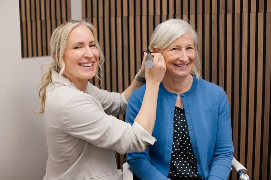 A woman with long blonde hair using an otoscope to examine an elderly woman's ear in a medical setting with wooden wall panels in the background.