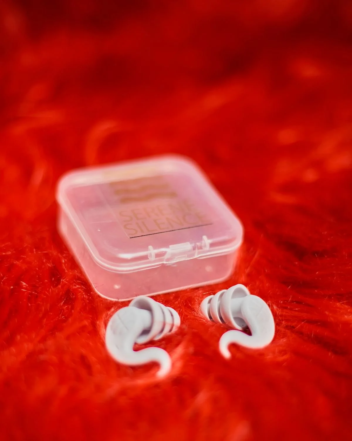 White in-ear headphones with a small clear plastic case on a red furry surface.