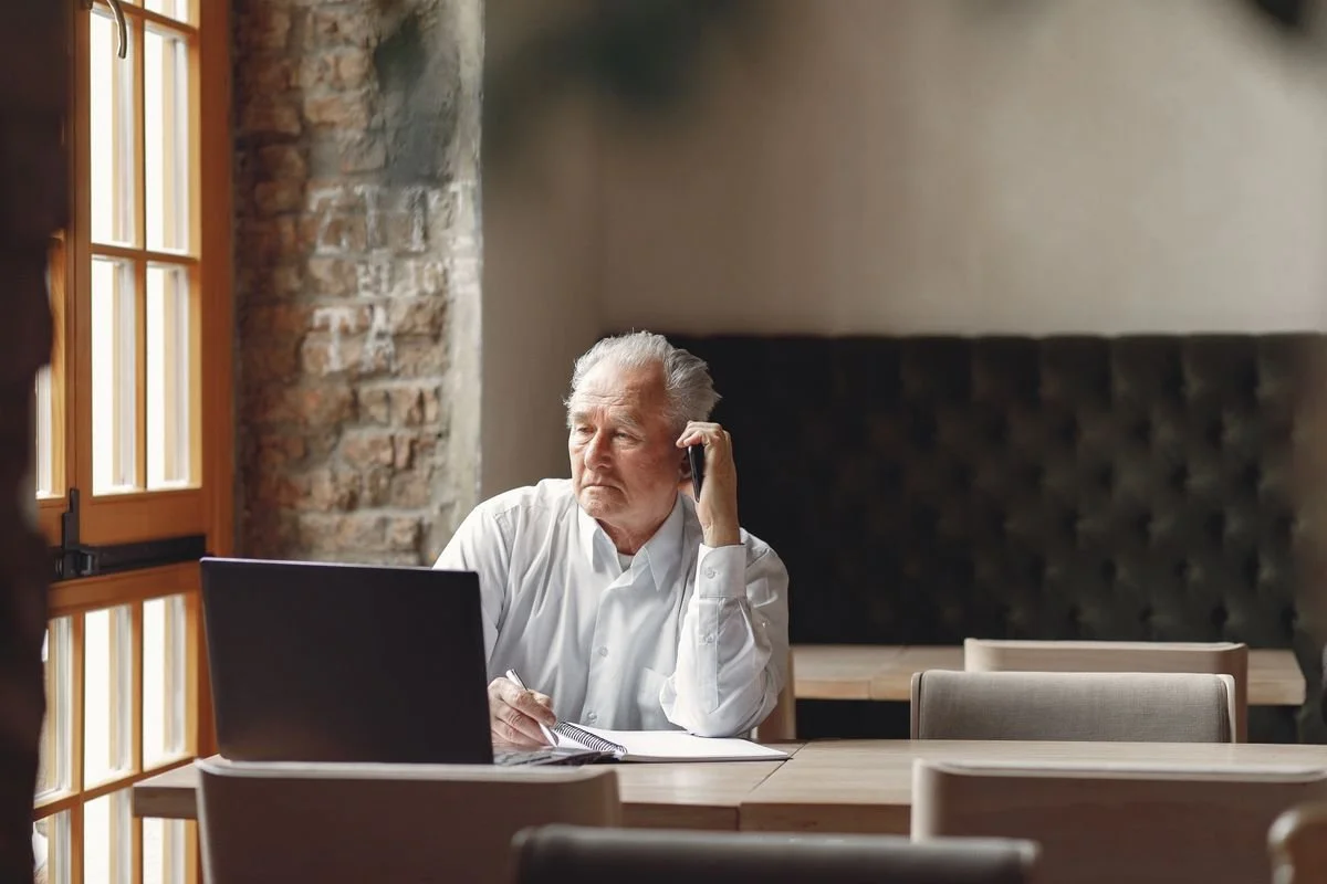 An elderly man in a white shirt sitting at a wooden table, working with a laptop, talking on the phone, inside a cozy room with brick walls and large windows.