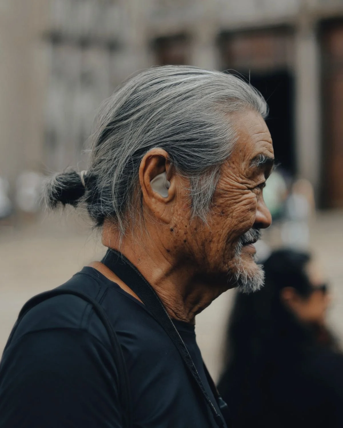 Profile view of an elderly man with long gray hair tied back, wearing a black shirt, standing outdoors with a blurred background.