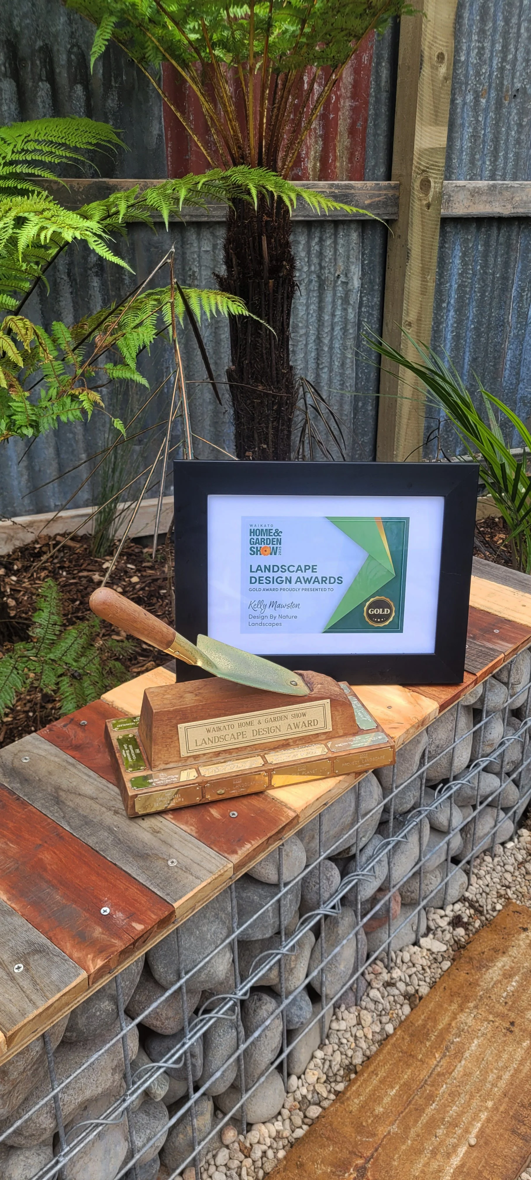 A framed certificate and a wooden award plaque with a gardening trowel on top, displayed on a garden table. The certificate is from the Waikato Home & Garden Show Landscape Design Awards, awarded to Kelly Mawston. The background includes a fern plant and a corrugated metal fence.