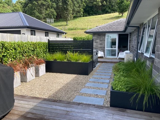 View of a backyard patio with stepping stones, raised garden beds, and outdoor seating area next to a house, with a neighboring house and green hill in the background.