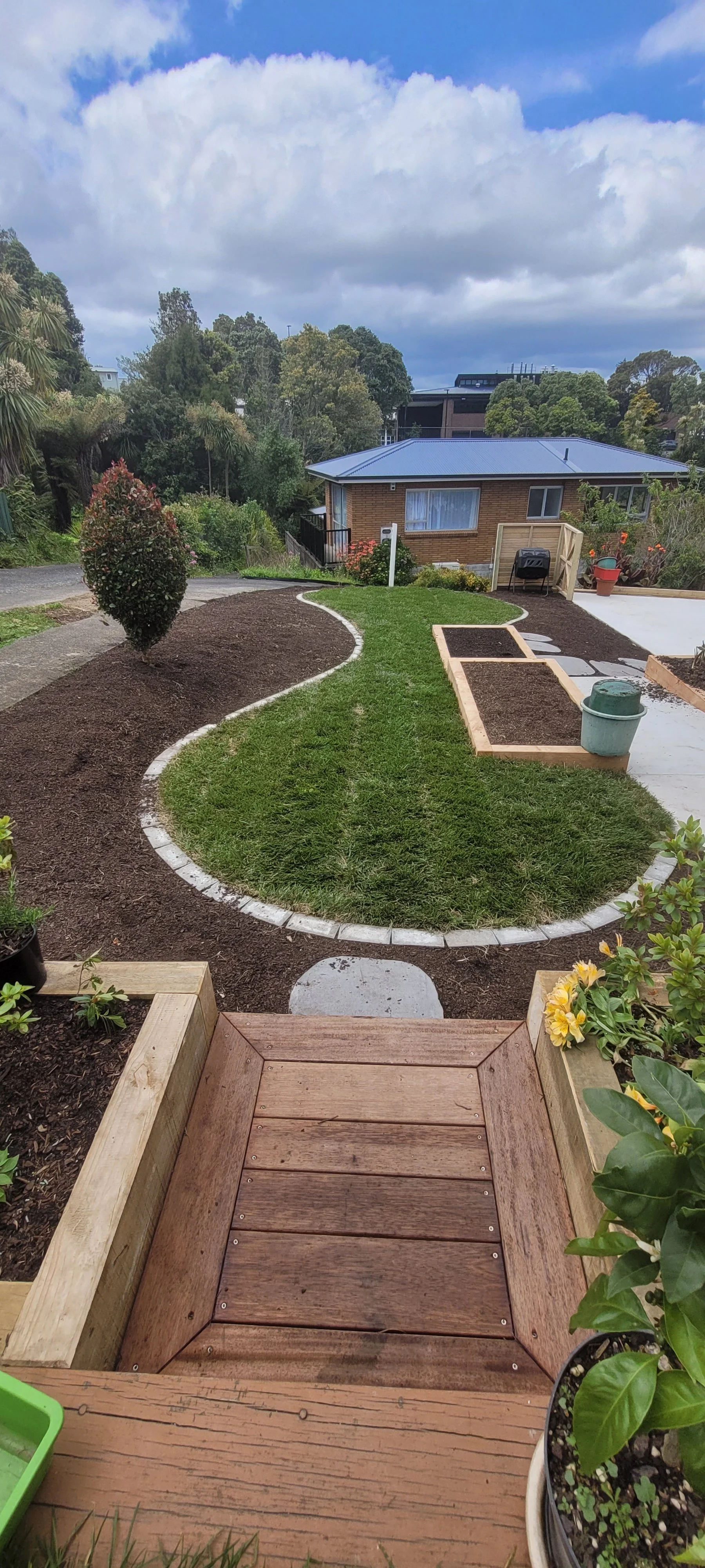 View of a backyard garden with a curved grassy path, landscaped flower beds, potted plants, and a small wooden deck in the foreground. A brick house with large windows is in the background under a cloudy sky.