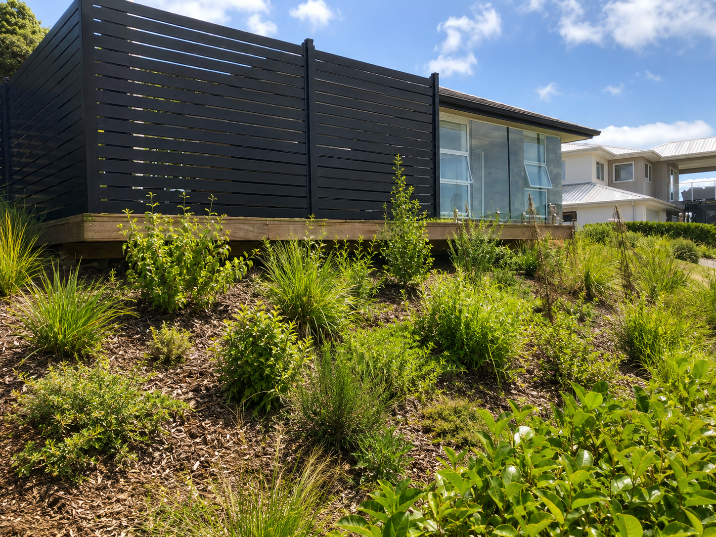A modern house with large windows, a wooden deck, and black horizontal slats fence, surrounded by lush green plants and shrubs in a landscaped garden on a sunny day.