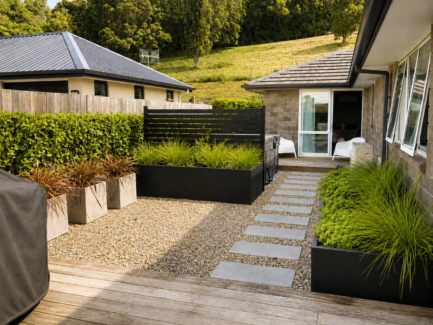 Modern backyard patio with potted plants, a gravel section, a wooden deck, lush green hedge fencing, and a sliding glass door leading into a house. There are two white outdoor chairs on the deck and a hillside with trees in the background.