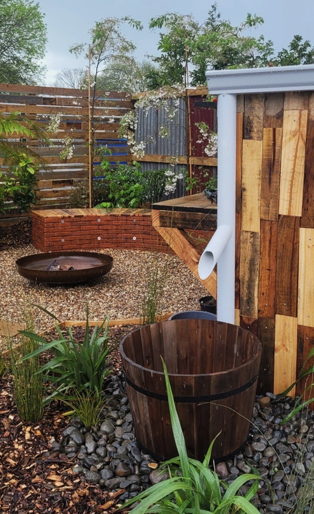 A backyard garden with a wooden shed, a fire pit, plants, and a gravel path, enclosed by a wooden fence.