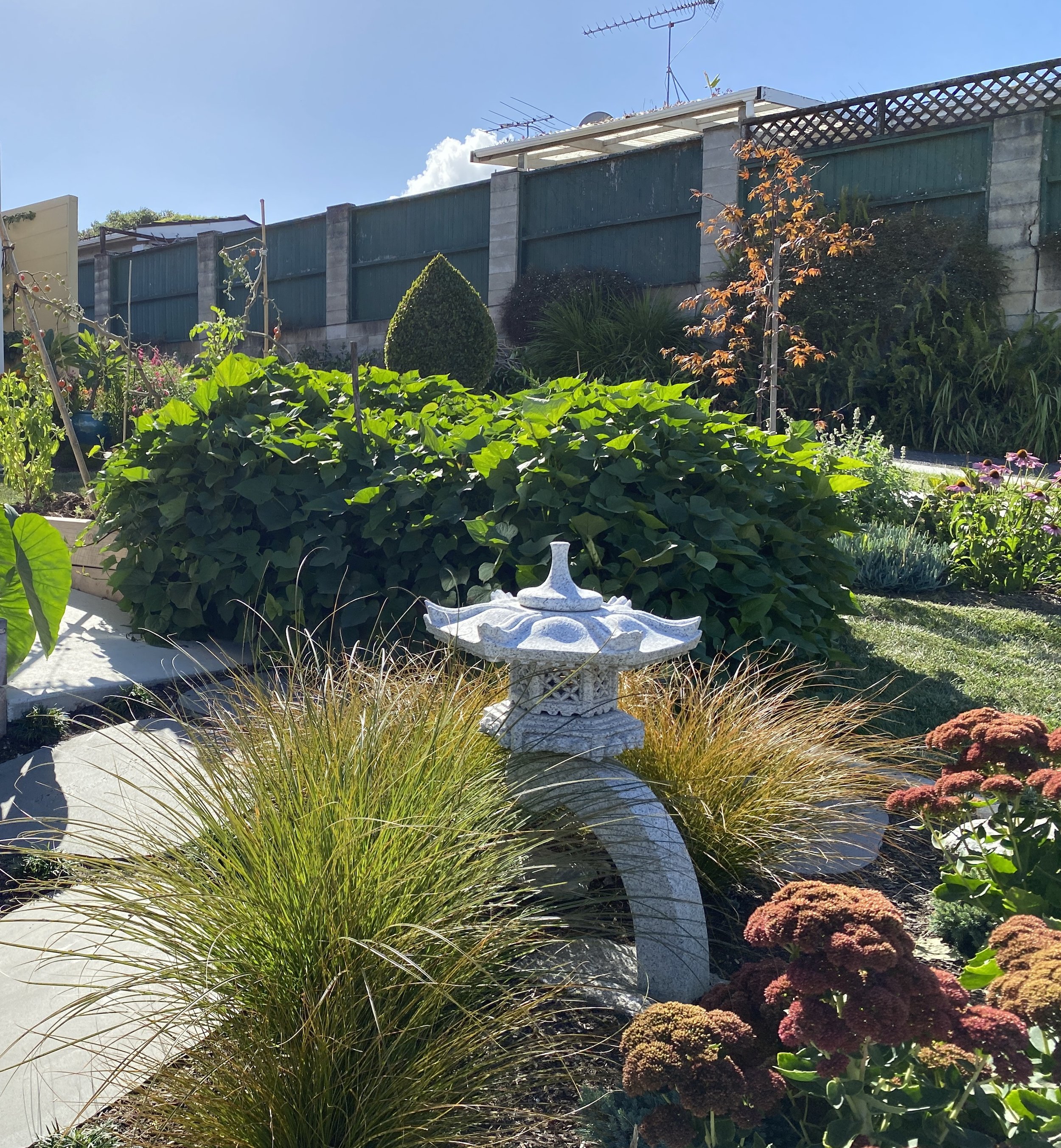 A lush garden with green leafy plants in the foreground, ornamental bushes with red and orange flowers, and a blue fence with stone pillars in the background. A white stone birdbath or garden ornament is also visible.