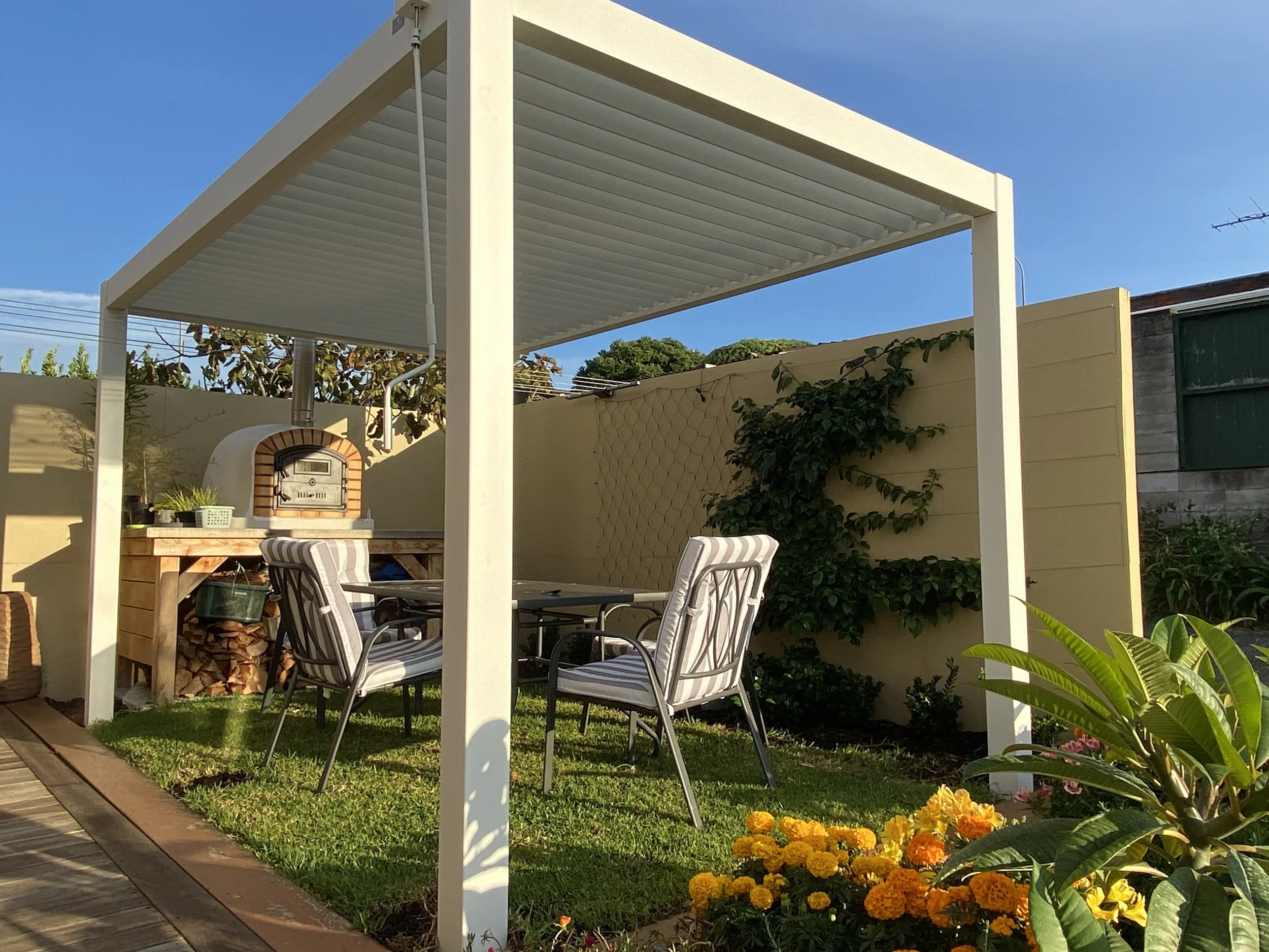 An outdoor patio area with a white pergola overhead, surrounded by a garden with flowering plants and a yellow fence, featuring patio chairs and a brick oven.