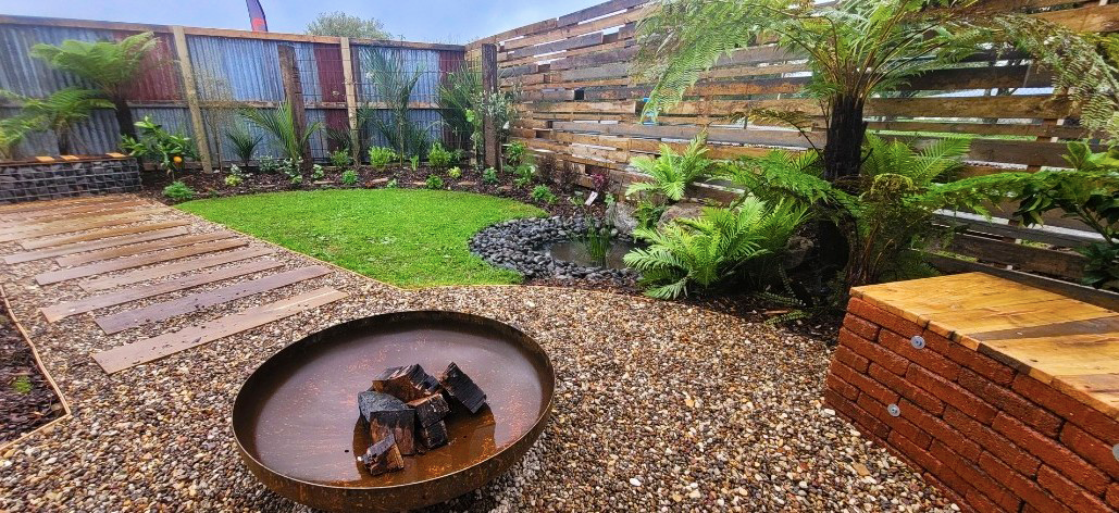 A backyard garden with a circular water feature containing wooden logs, a pathway with stone slabs, lush green plants, a small patch of grass, and a wooden and brick structure along a wooden fence.