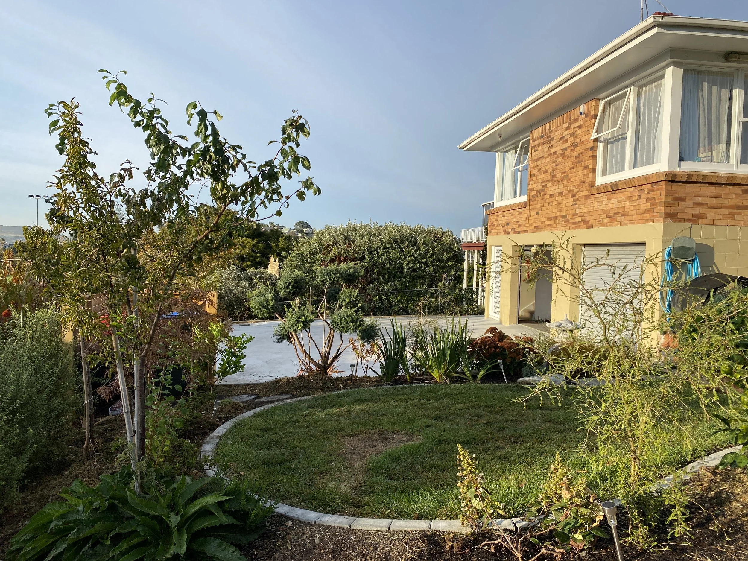 A backyard with a variety of trees, shrubs, and plants, a brick house with open windows, a grass lawn, and garden lights.