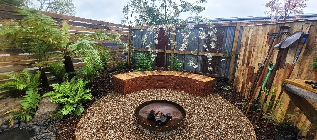 A small backyard garden with a gravel circle in the center around a fire pit, a curved brick bench, green plants, a wooden fence, and gardening tools leaning against the fence.