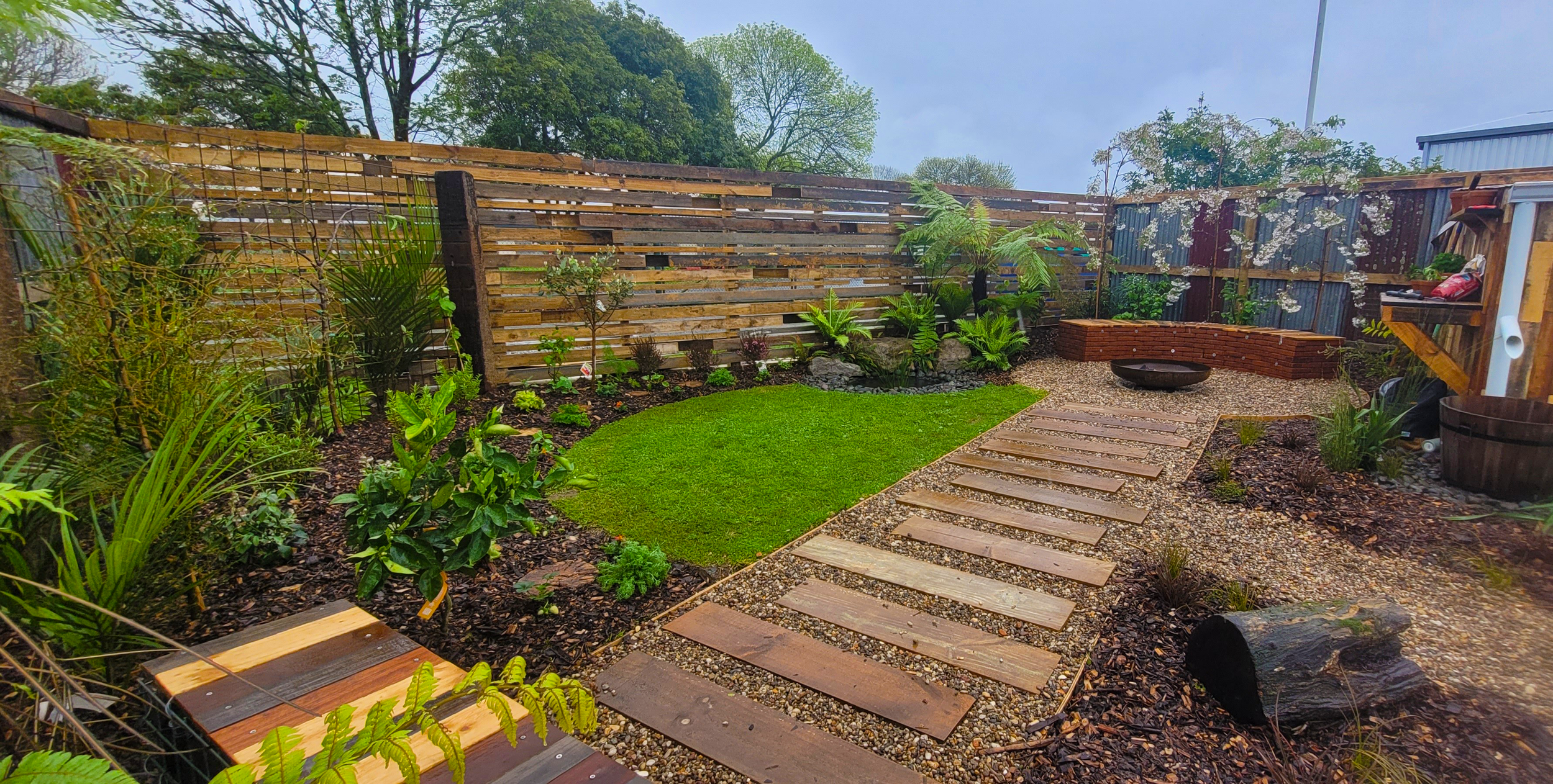 A backyard garden with a wooden privacy fence, plants, small lawn, stone pathway, and outdoor storage on the right.