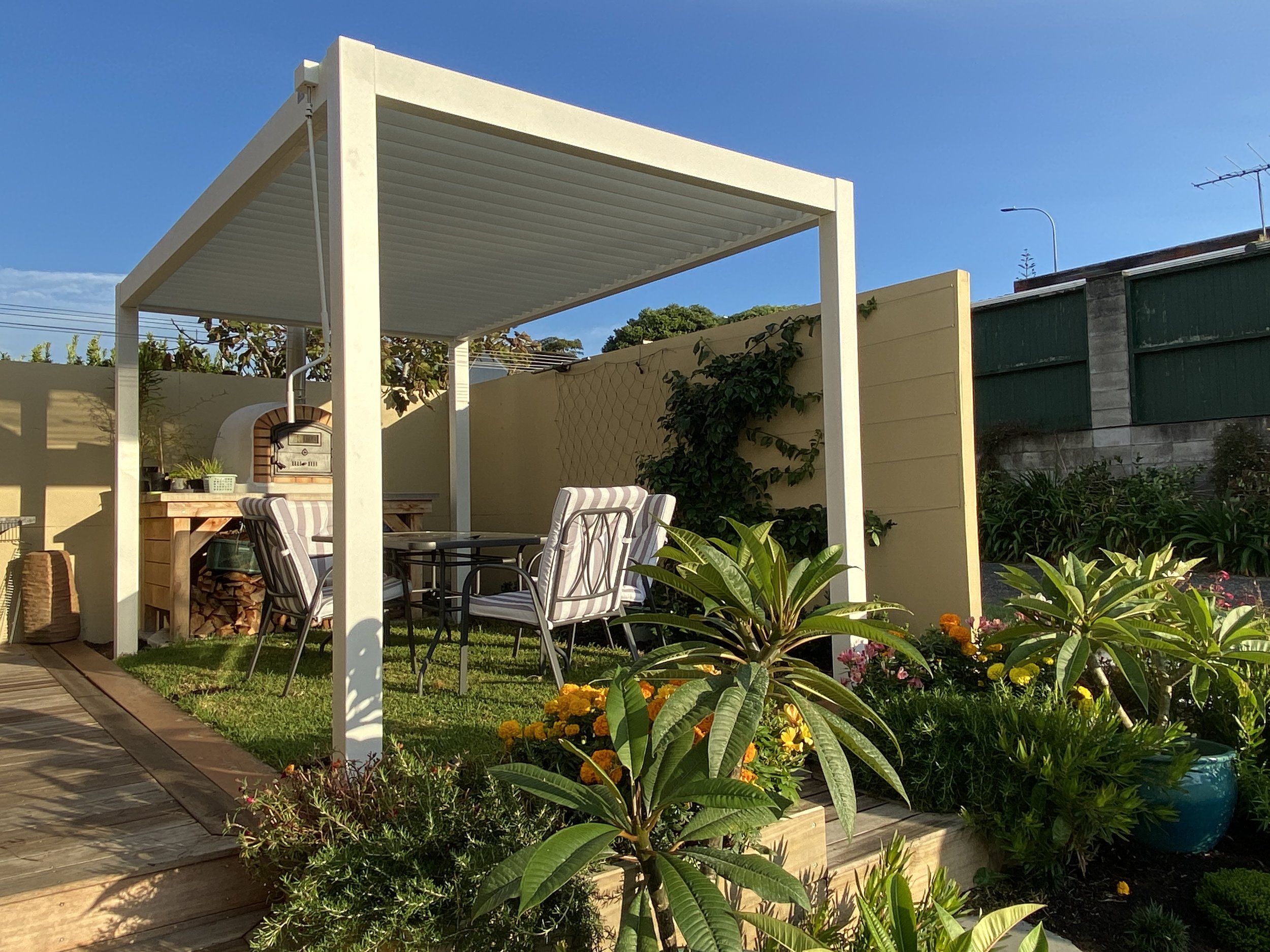A backyard patio with a white pergola, outdoor dining table, chairs, and colorful flower garden under a clear blue sky.