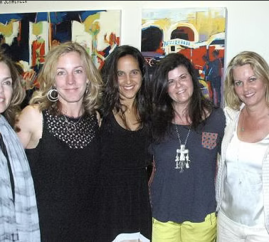 A group of five women standing together outdoors, with colorful abstract paintings in the background.