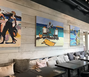 Three colorful beach-themed paintings hung on a concrete wall inside a cafe or restaurant with a cushioned seating area and black tables.