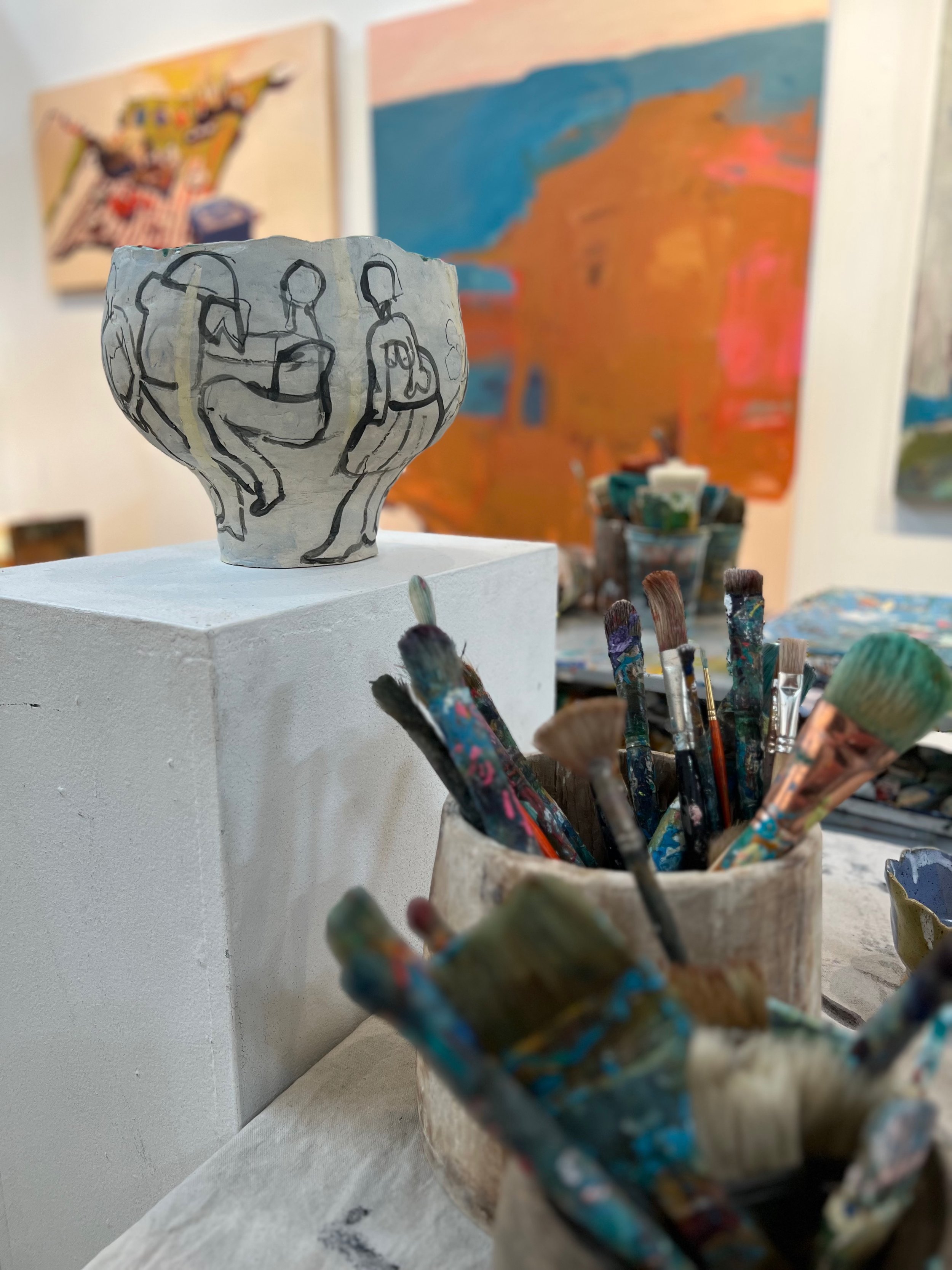 A ceramic bowl with black line drawings of people on it, placed on a white pedestal in an art studio. In the background, colorful abstract paintings hang on the wall, and paintbrushes in a ceramic container are visible in the foreground.