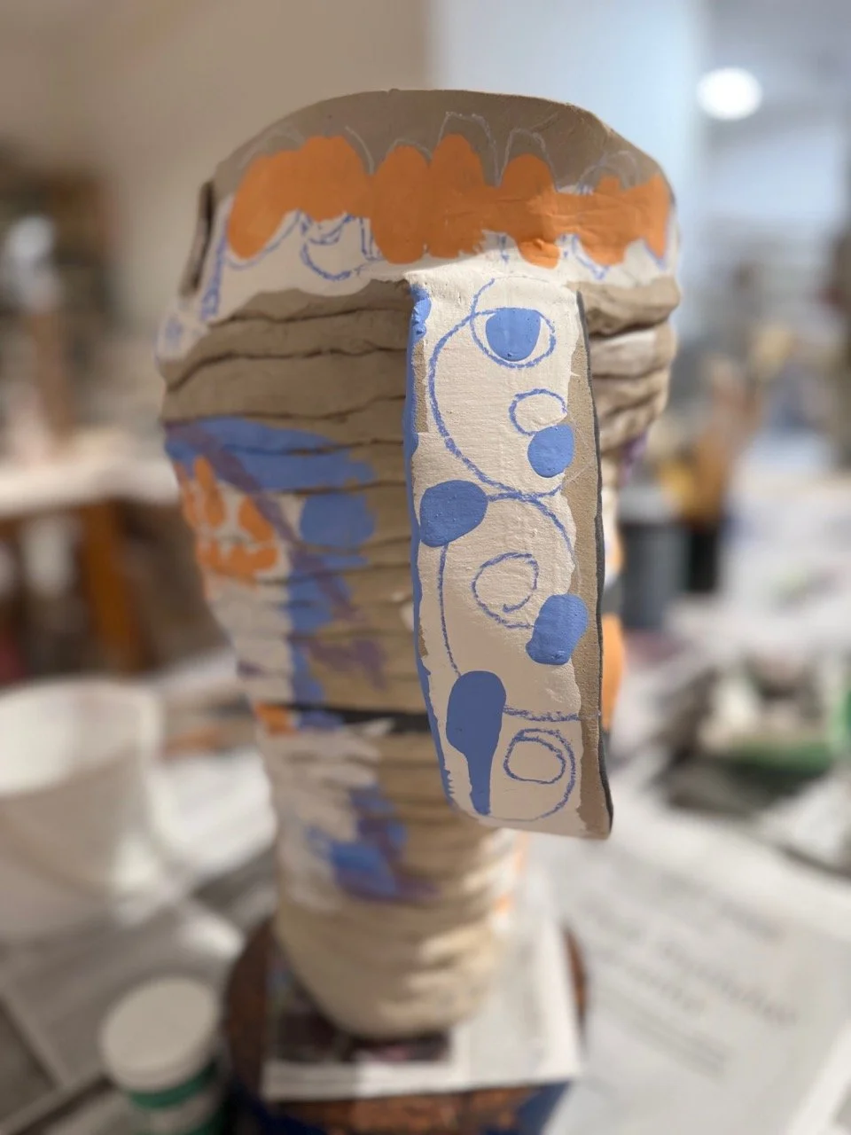 A handcrafted sculpture resembling a face, made from twisted paper or cardboard with painted decorations, including orange and blue designs, in an artistic or craft setting.