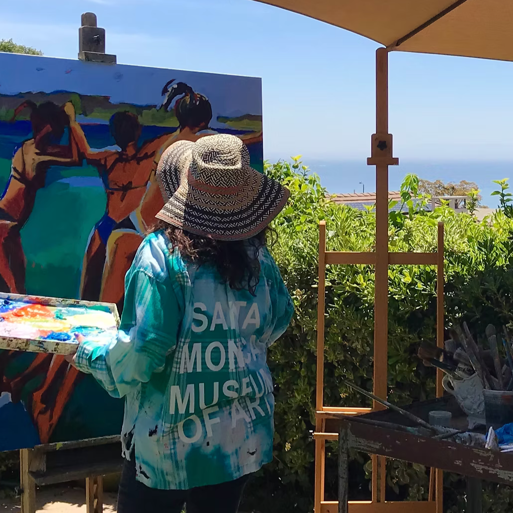 An artist wearing a sun hat and a jacket labeled 'SANTA MONICA MUSEUM OF ART' paints an outdoor landscape with a mostly blue sky, greenery, and water in the background. The artist's palette with various paint colors is visible, and the scene is set under a tent or canopy.