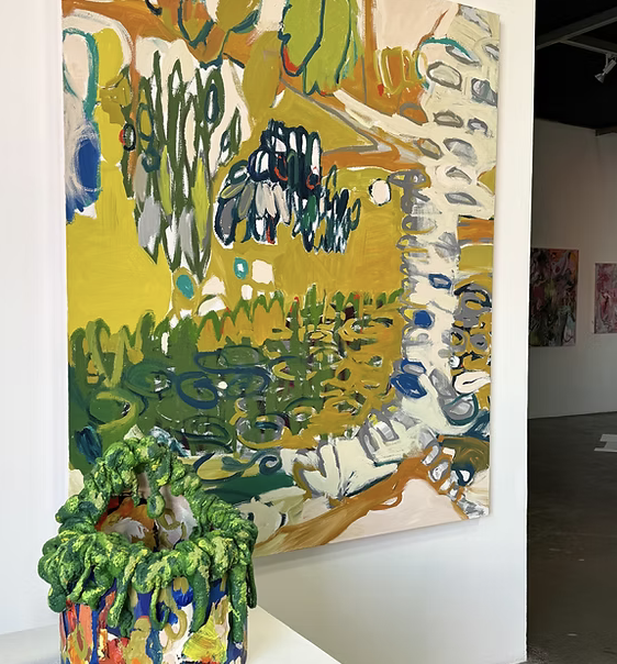 Colorful abstract painting on a wall featuring green, yellow, blue, white, and orange brushstrokes, with a small plush doll or sculpture in the foreground that is green with a multicolored base.