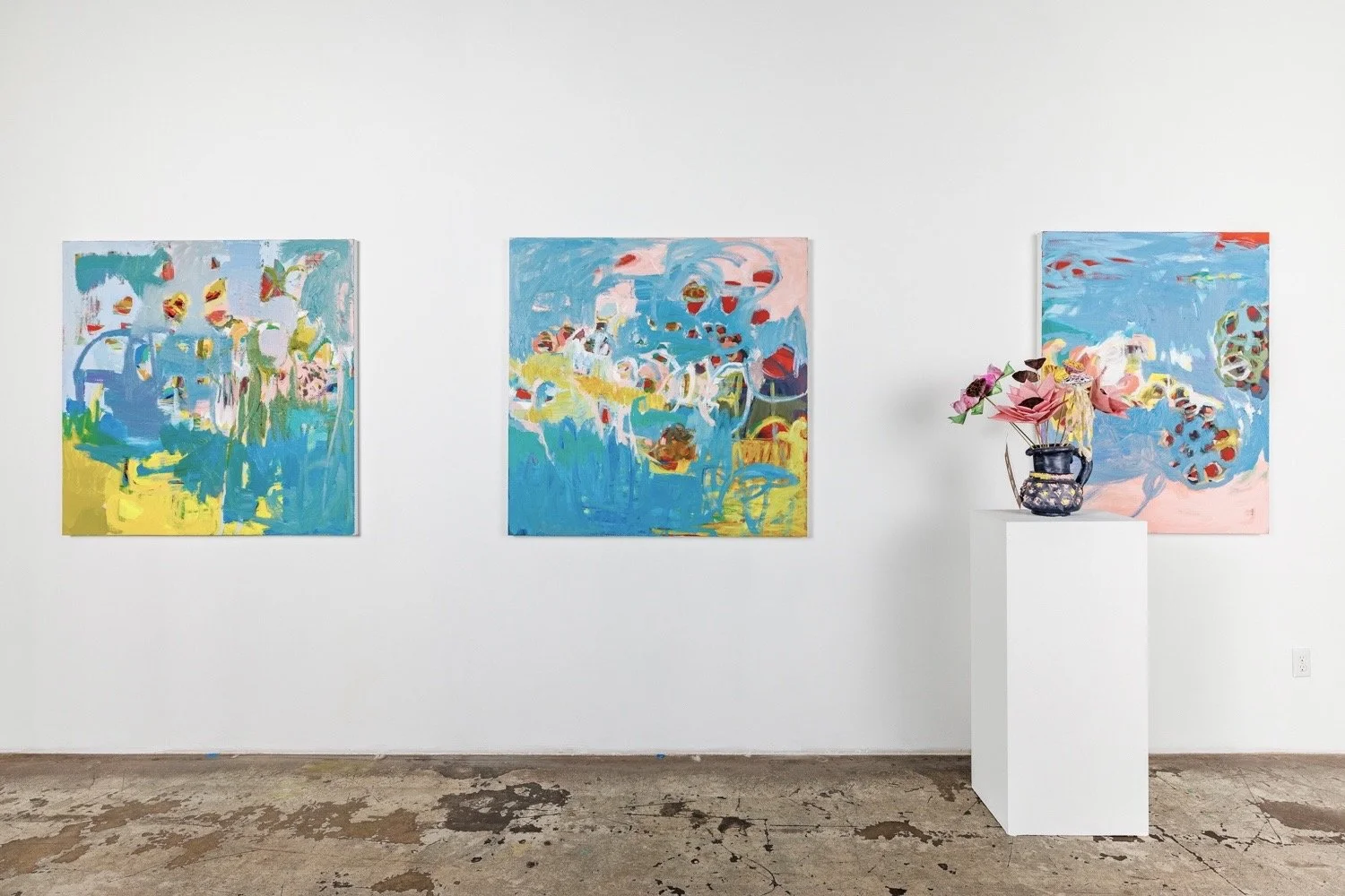 An art gallery with three colorful abstract paintings on a white wall and a flower arrangement in a dark vase on a white pedestal.