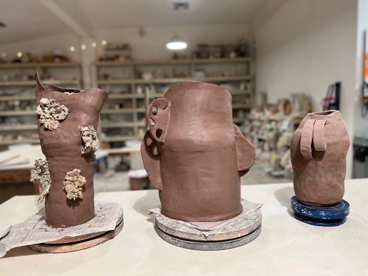 Three ceramic vessels in various stages of completion on a shelf in an art studio, with shelves filled with art supplies in the background.