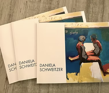 Three artist's books titled "Daniela Schweitzer" with one open showing a colorful painting of two people with gray hair sitting and looking at a book or paper.