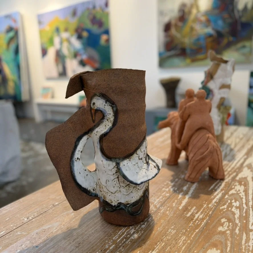 Ceramic swan sculpture with a dark brown hat and scarf in an art gallery, with abstract paintings and sculptures in the background.