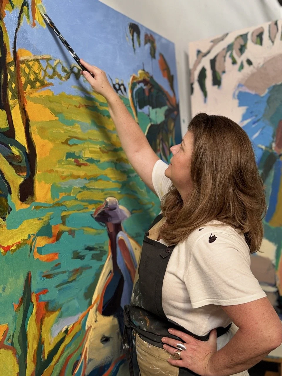 A woman with brown hair wearing a white T-shirt and black apron painting a colorful landscape on a large canvas. She is using a brush and has her right hand on her hip, smiling as she works.